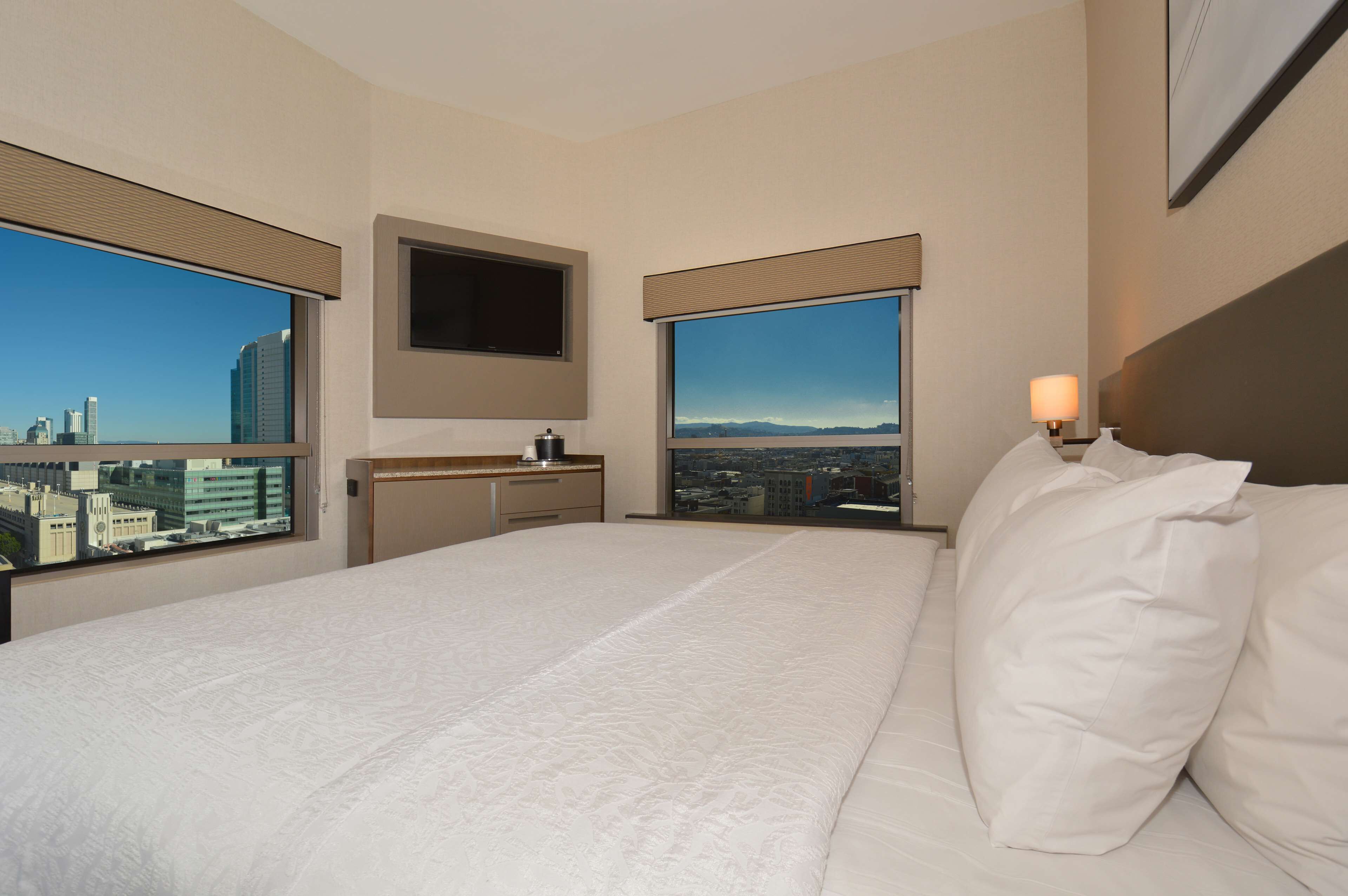 A hotel room with a large bed, a TV mounted on the wall, a window with a city view, and a bedside table with a lamp.
