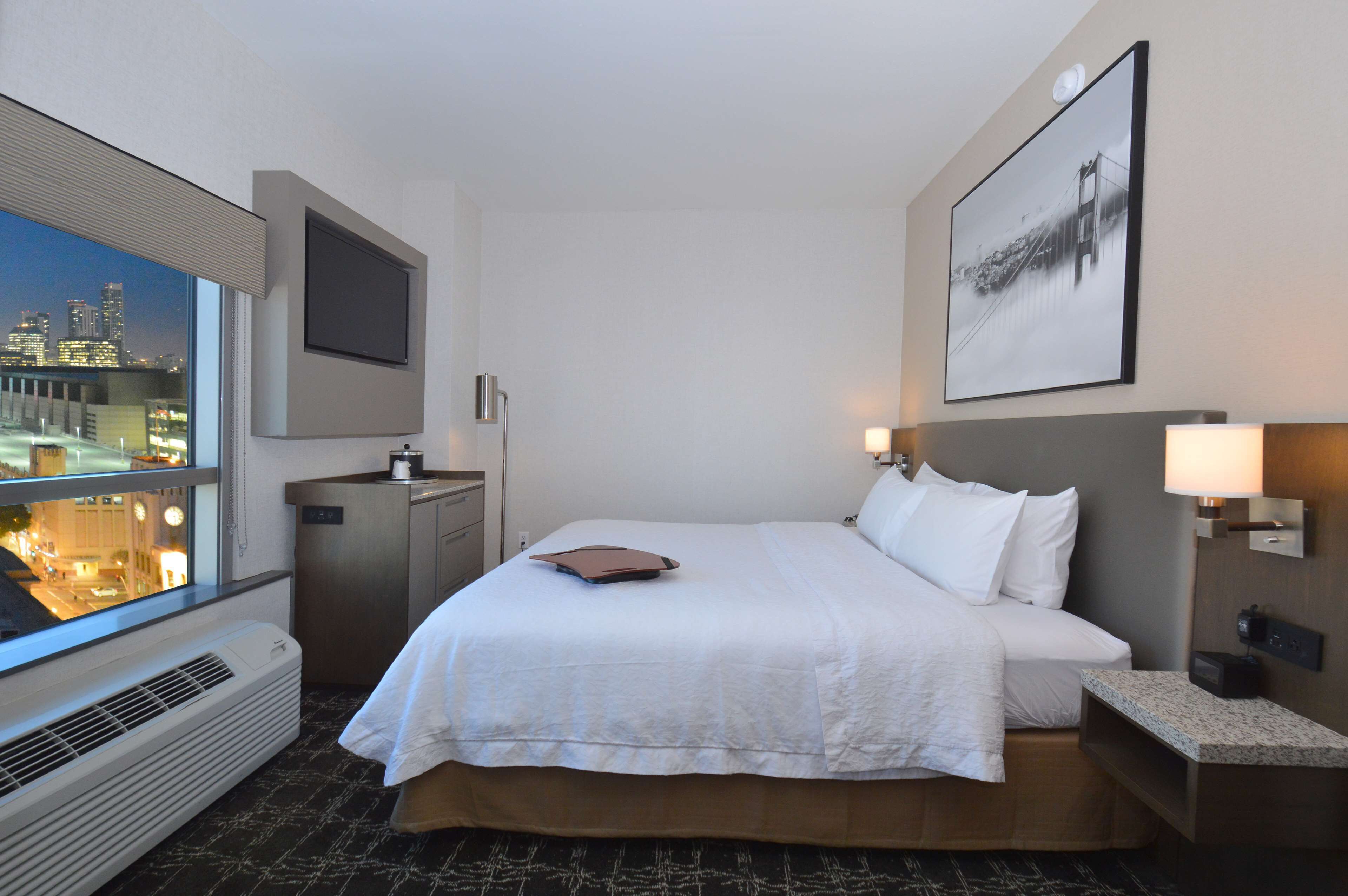 Habitación de hotel con una cama grande, un escritorio, una silla, un televisor montado en la pared, un ventanal con vista a la ciudad y un cuadro enmarcado en la pared.