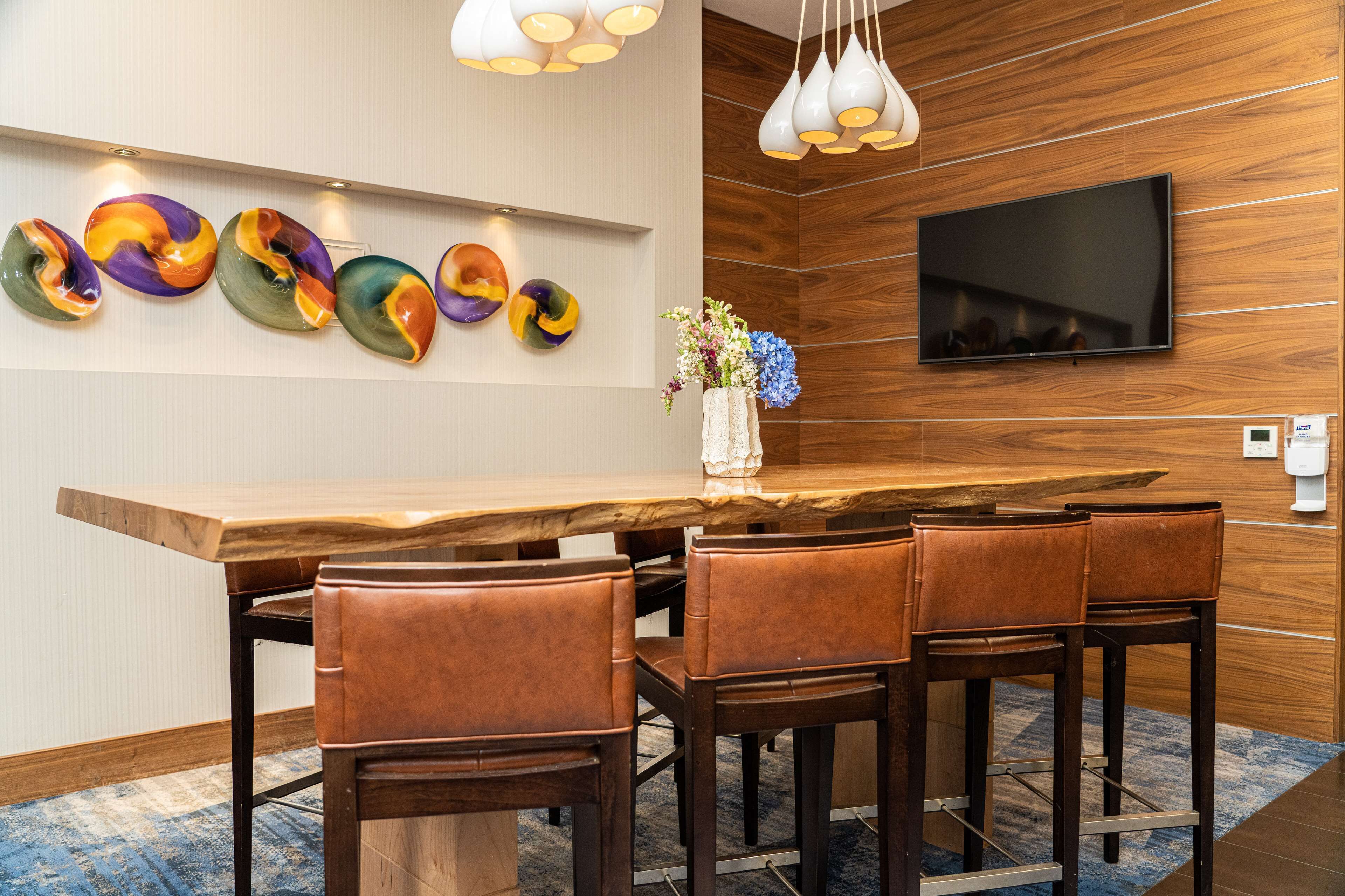 A modern dining area with a wooden table, leather chairs, a vase with flowers, and a flat-screen TV on the wall.
