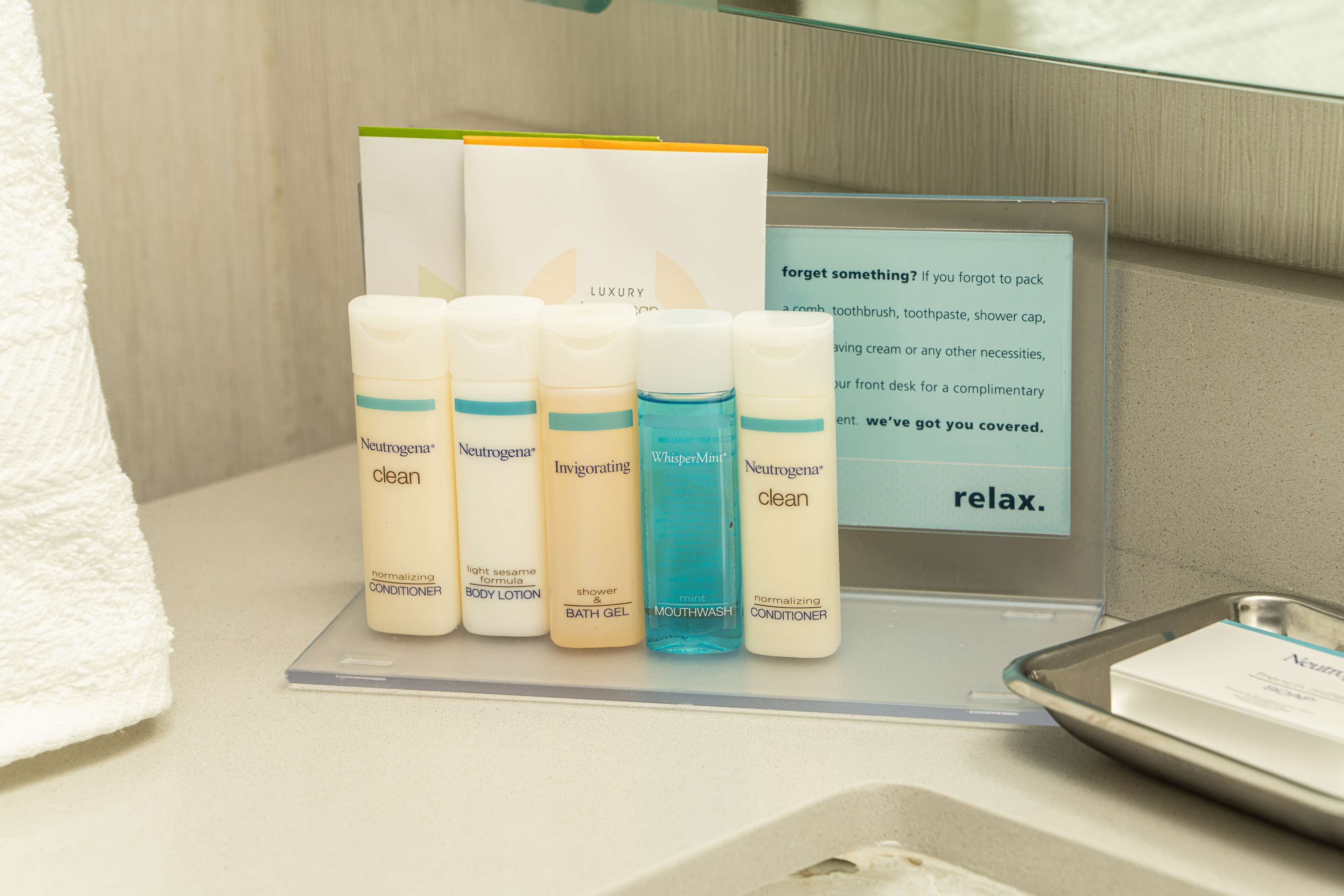 A bathroom counter with Neutrogena products, including body lotion, bath gel, and mouthwash. A sign offers complimentary items like a toothbrush and toothpaste.