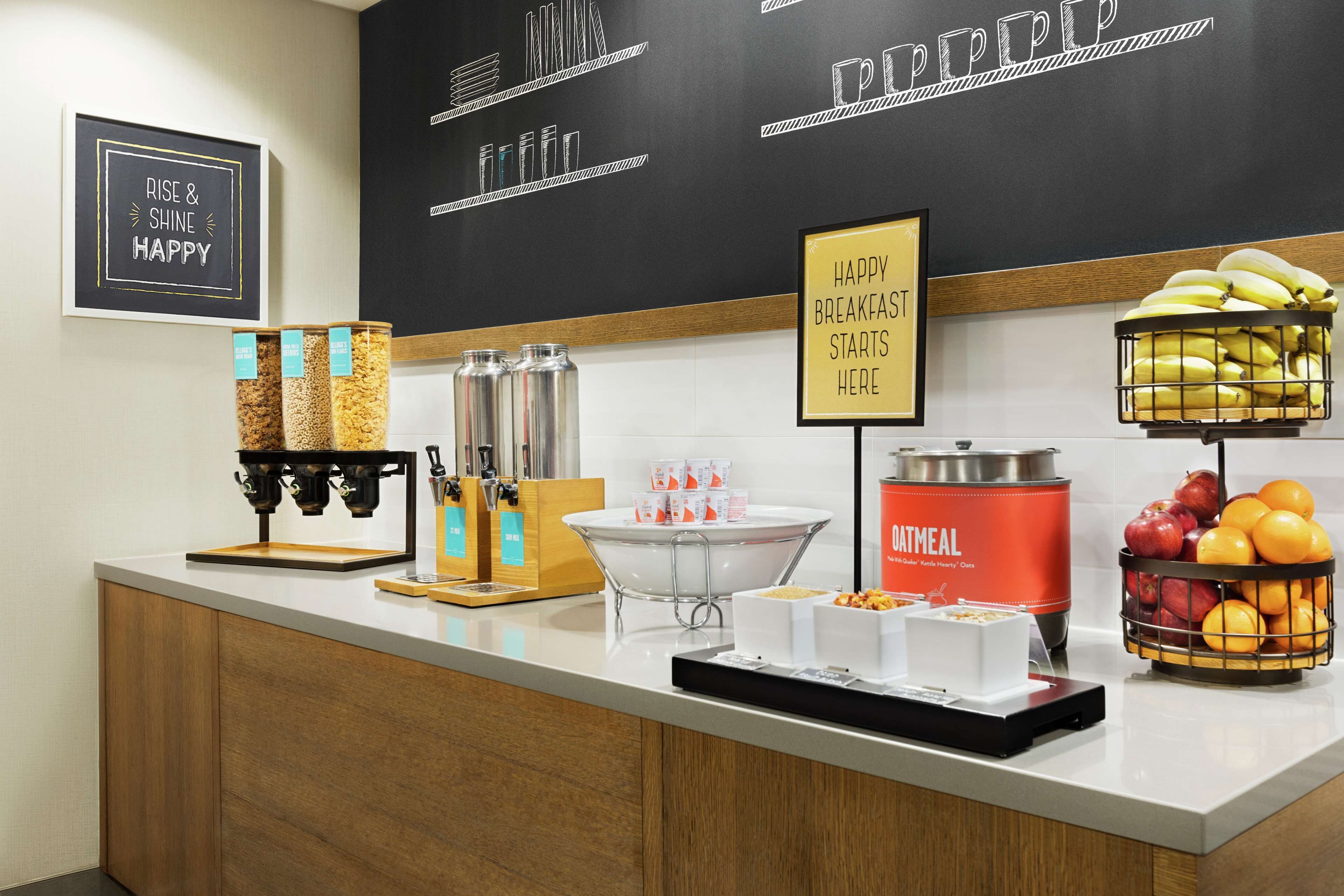 A breakfast bar with coffee machines, a fruit basket, and a sign indicating breakfast starts here.