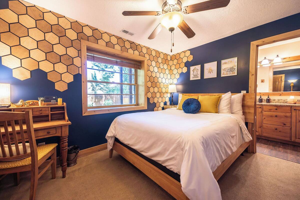 A bedroom with a wooden bed, a desk, and a chair. There is a window with a view of trees, a ceiling fan, and a mirror.