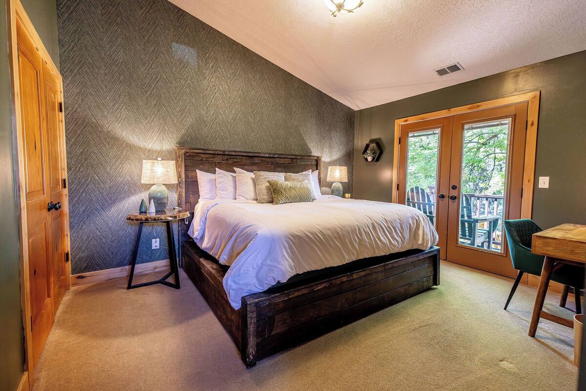 A bedroom with a large bed, a wooden headboard, a small table with a lamp, and a view through a door to an outdoor area.