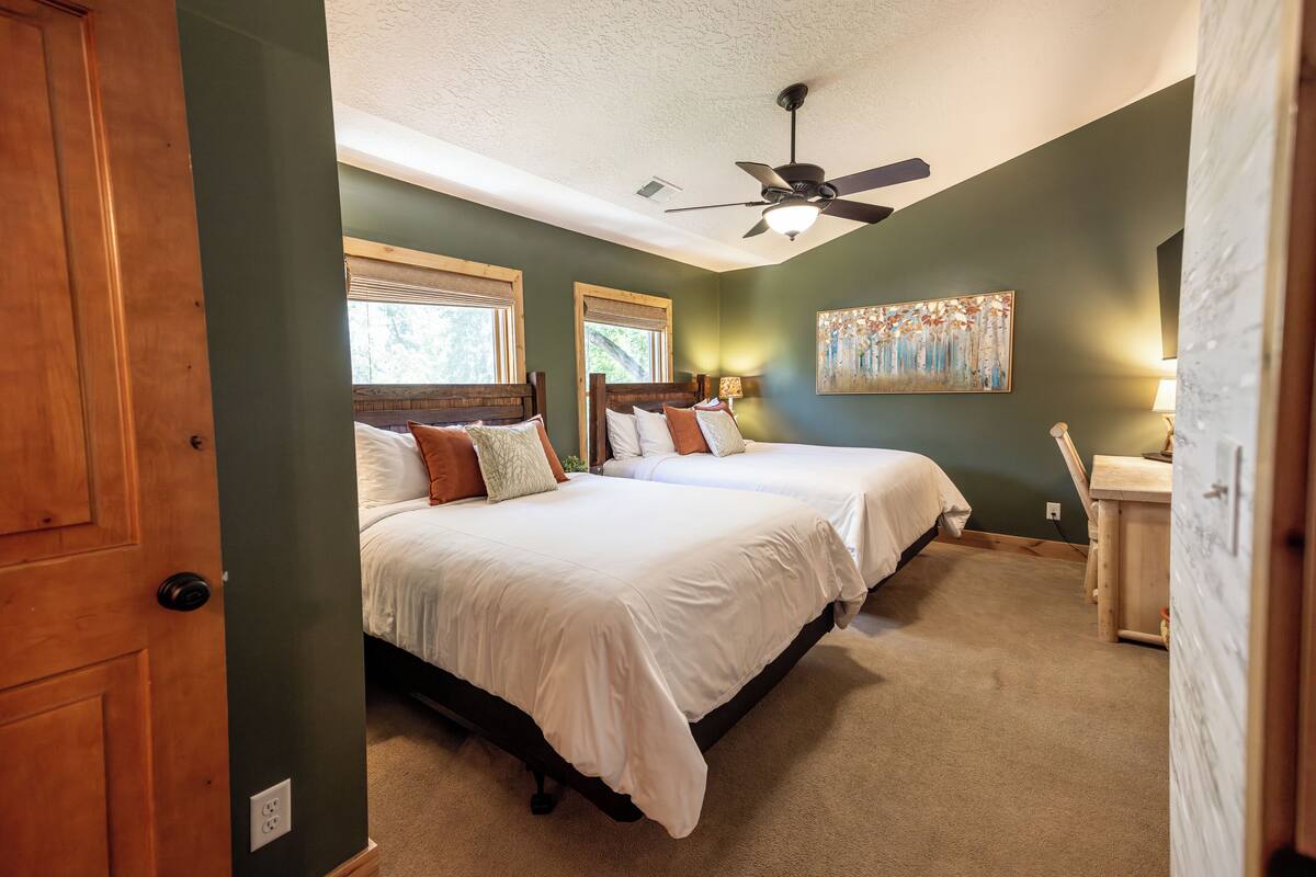 A bedroom with two beds, a ceiling fan, a painting, and a desk.