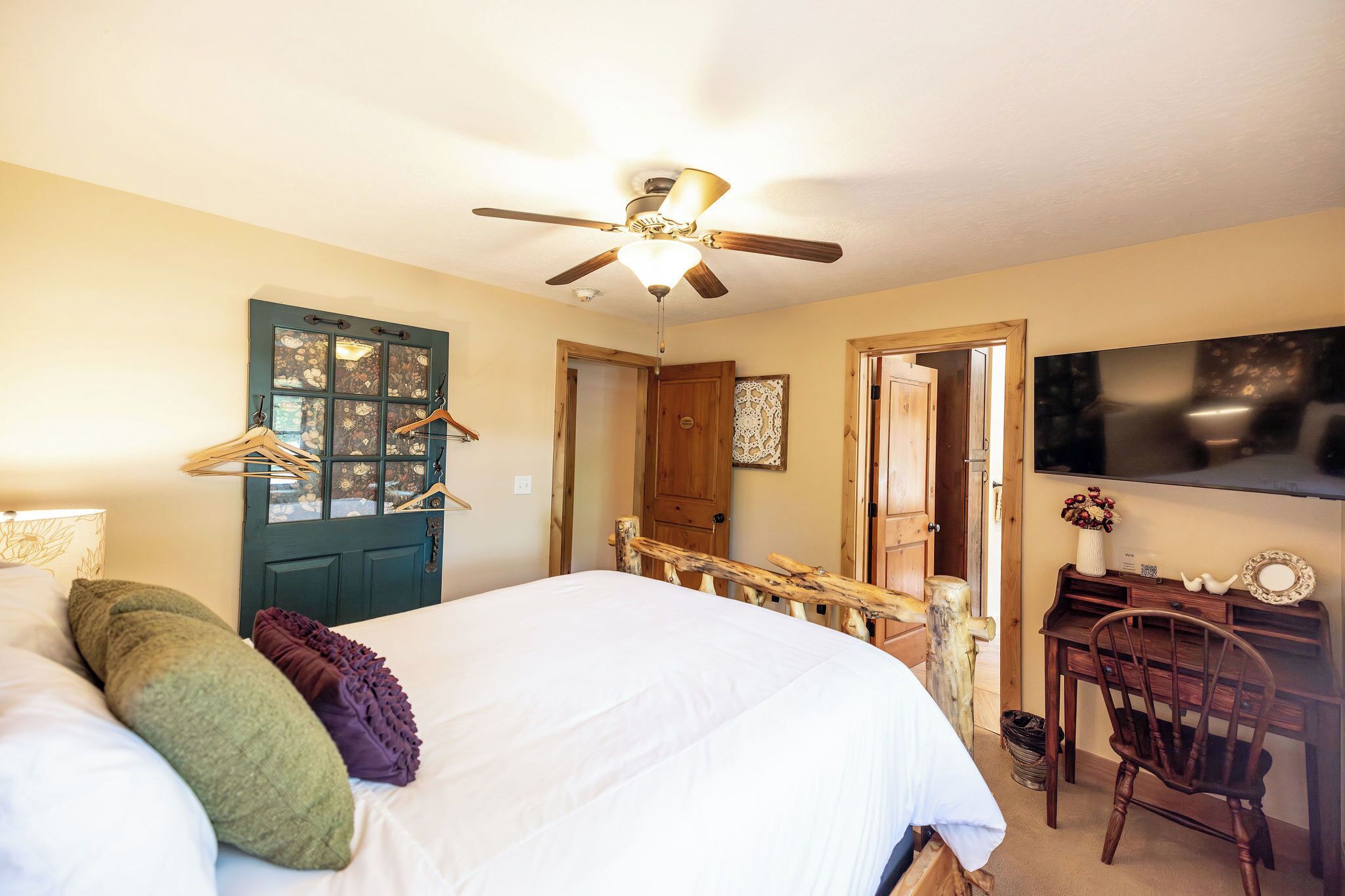 A bedroom with a bed, a desk, a chair, a ceiling fan, and a door.