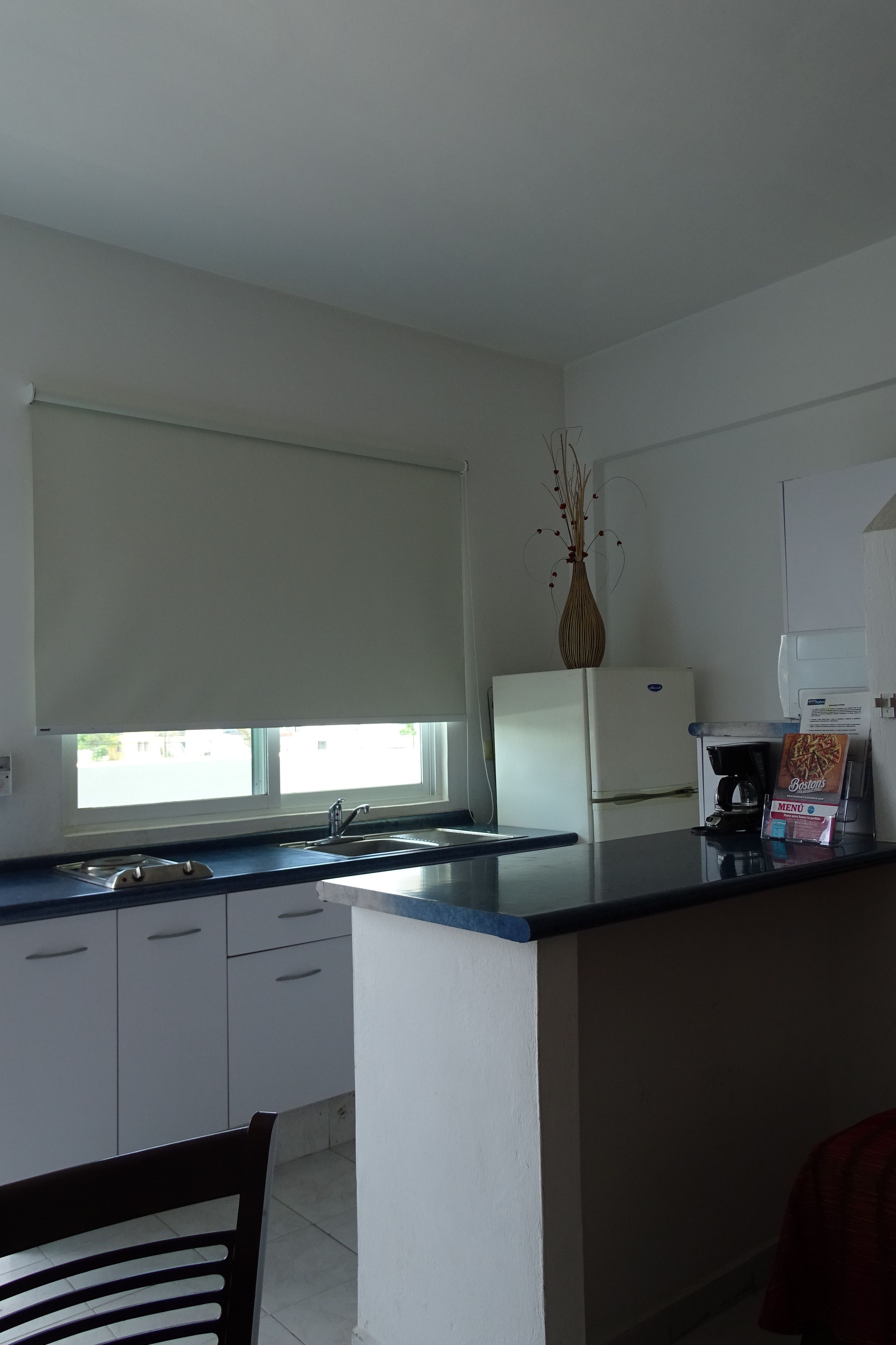 Cozinha com armários brancos, bancada preta, geladeira branca, máquina de café e um vaso com galhos no balcão.