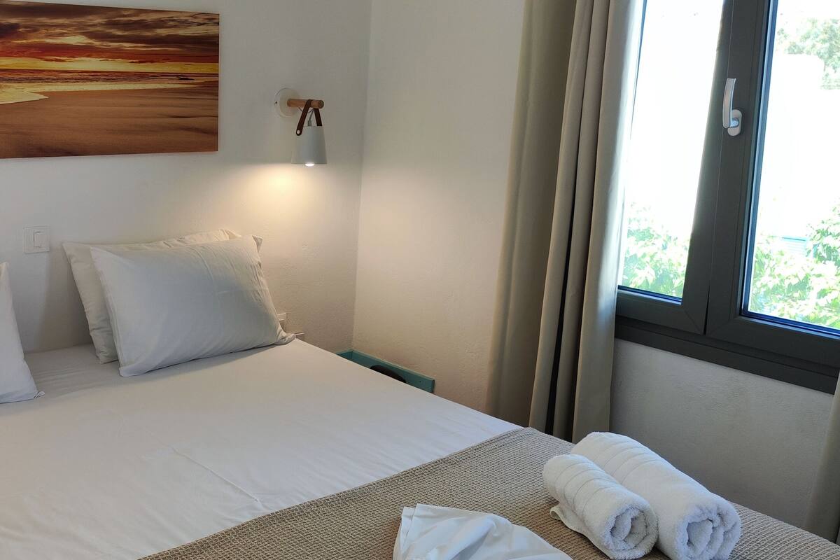 A neatly made bed with white linens and pillows, a painting of a sunset over the ocean on the wall, and a window with curtains.