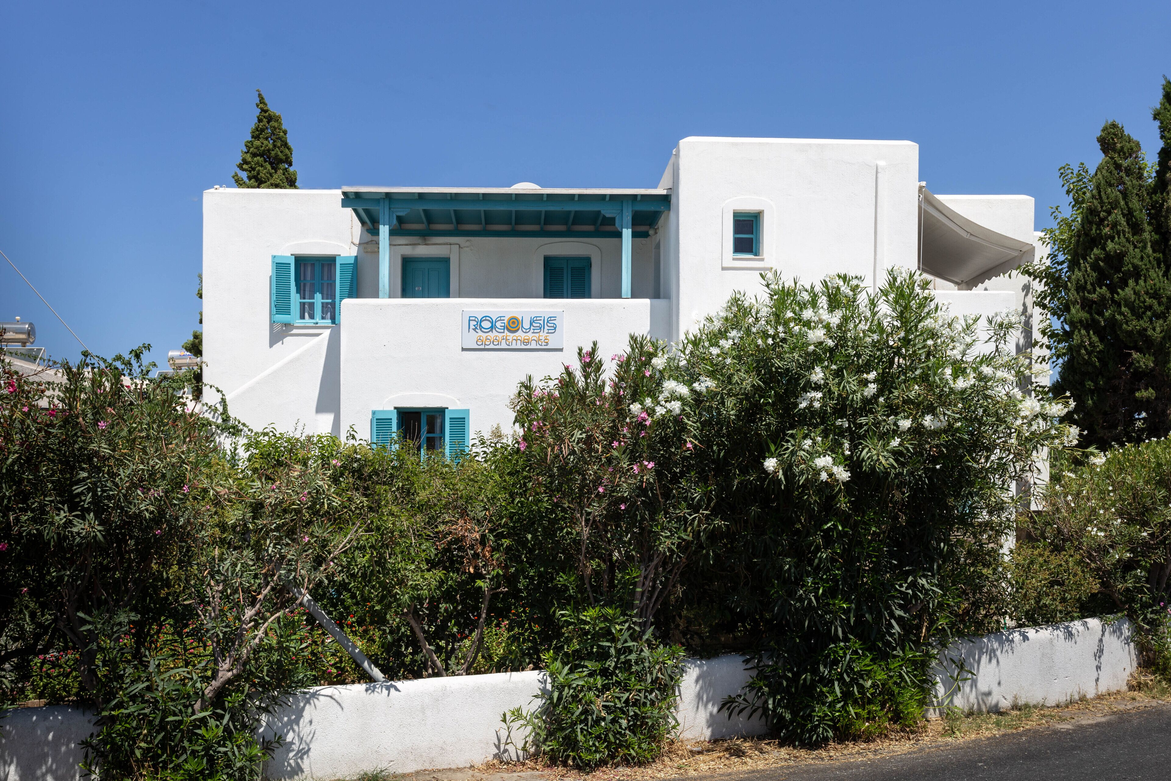 A white building with blue shutters and a sign reading "ROGOUSIS APARTMENTS".