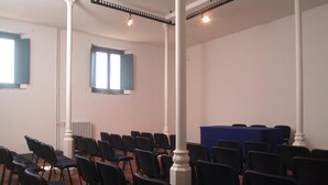 Una sala conferenze con file di sedie nere, colonne bianche e un tavolo blu.