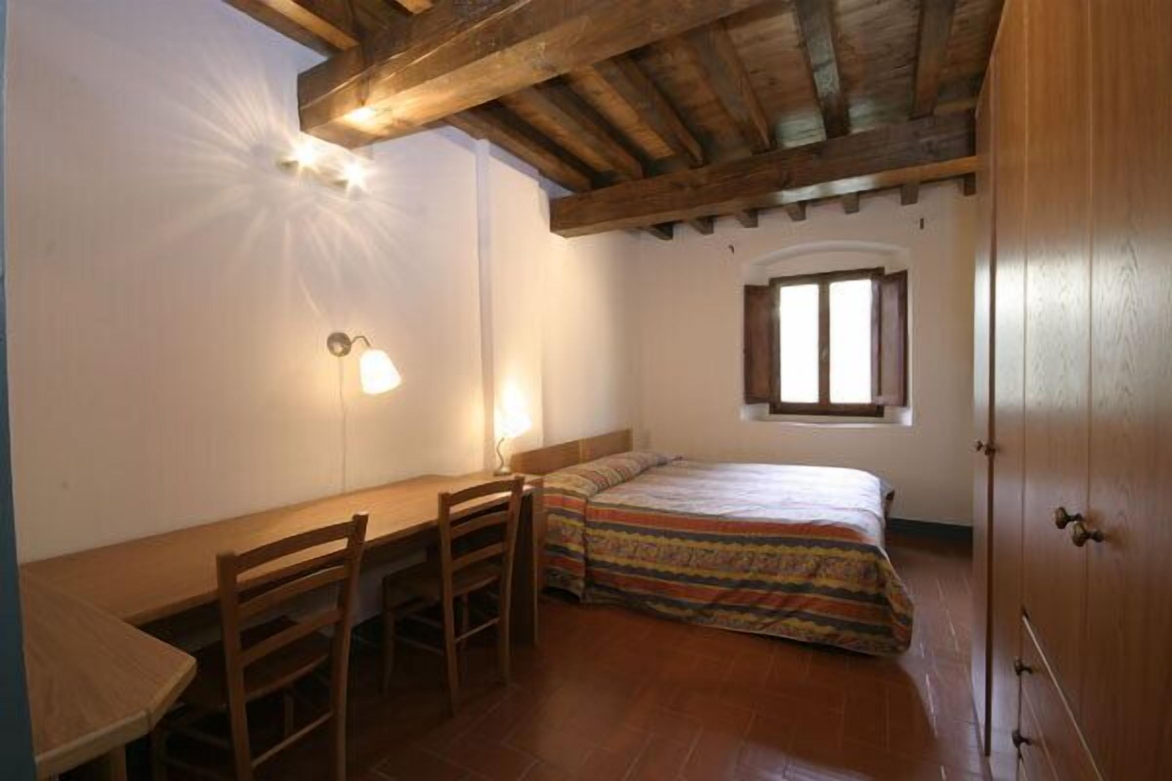 A small, cozy room with a wooden desk, a bed with a colorful blanket, a single wall-mounted light, and a window with blinds.