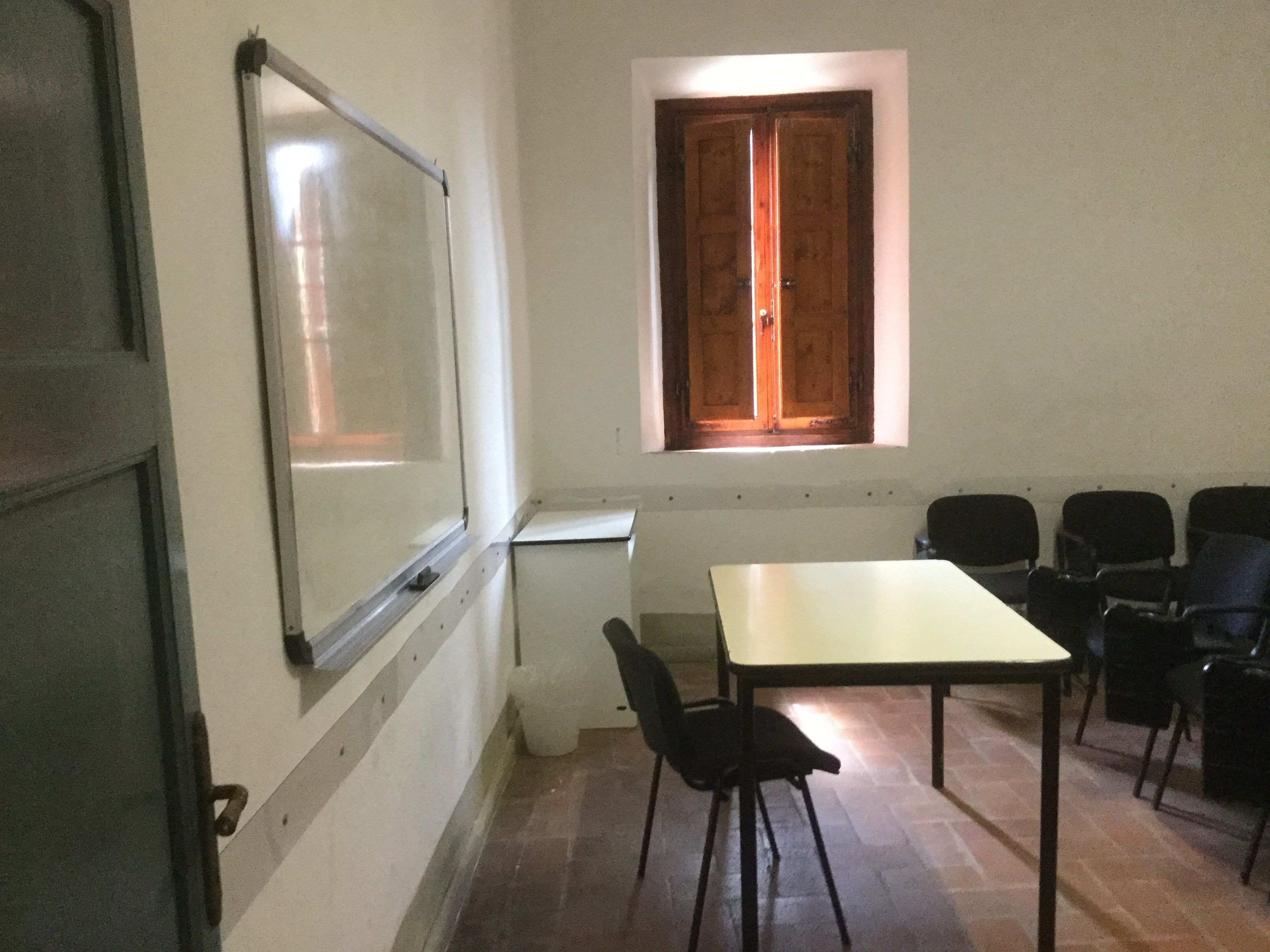 Une salle de conférence avec une table blanche, des chaises noires, un écran de projection et une fenêtre en bois.