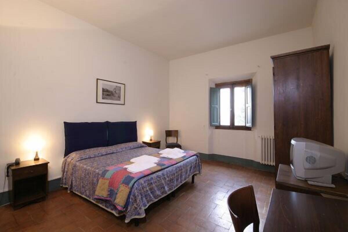 Un dormitorio con cama, lámparas de noche, un escritorio con impresora, una ventana y un radiador.