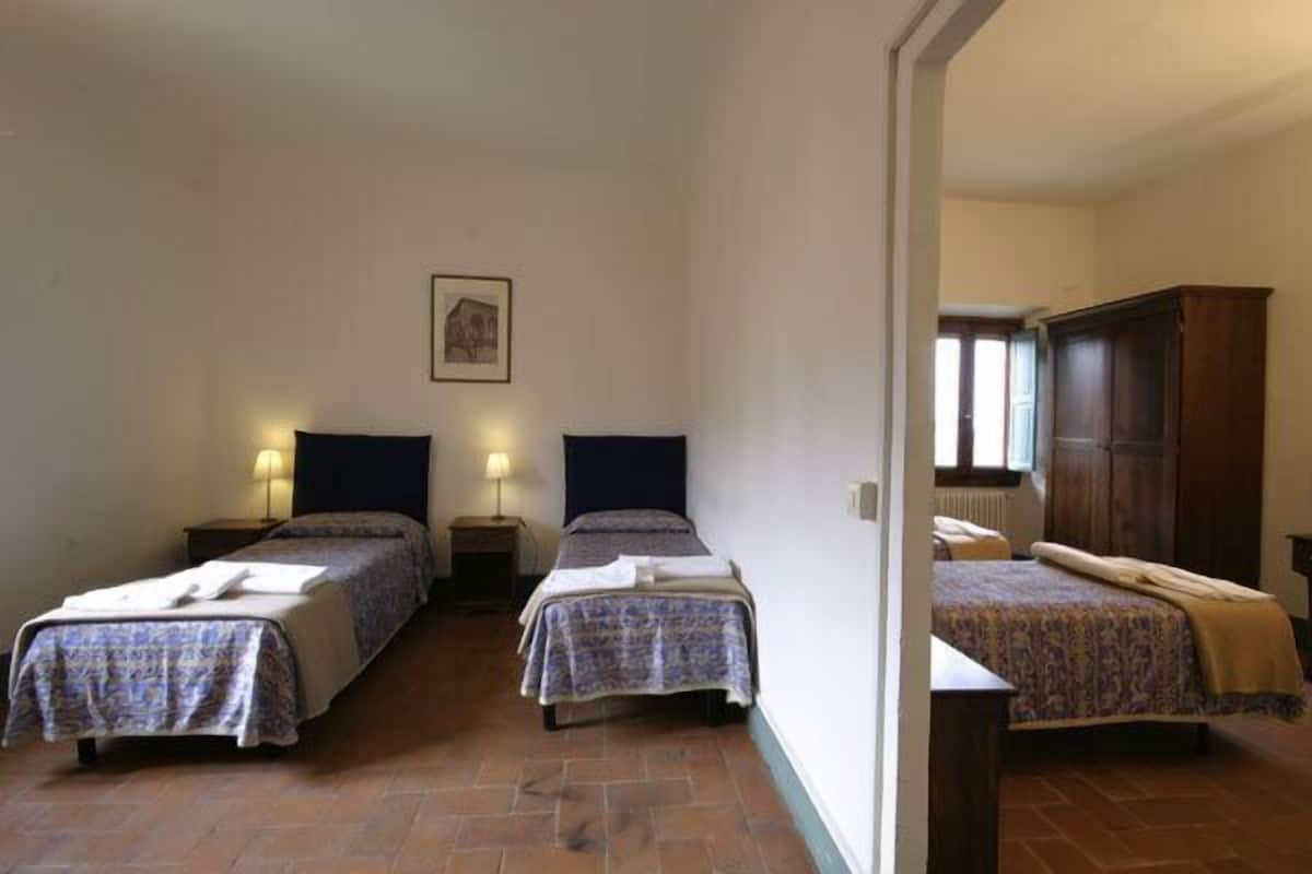 Una habitación con dos camas, cada una con cabecera y mesitas de noche. Hay un cuadro enmarcado en la pared y una ventana con cortinas.