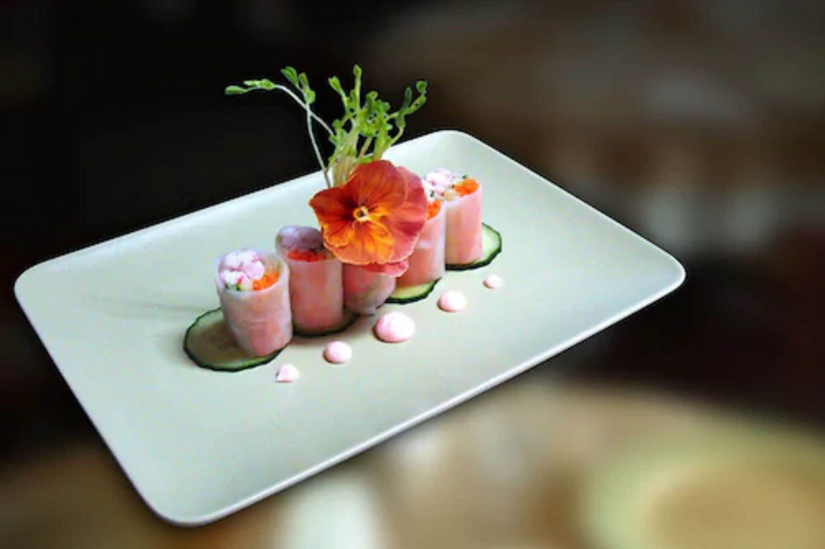 A rectangular plate with a food arrangement featuring pink and white elements, possibly sushi, garnished with a flower and green leaves.