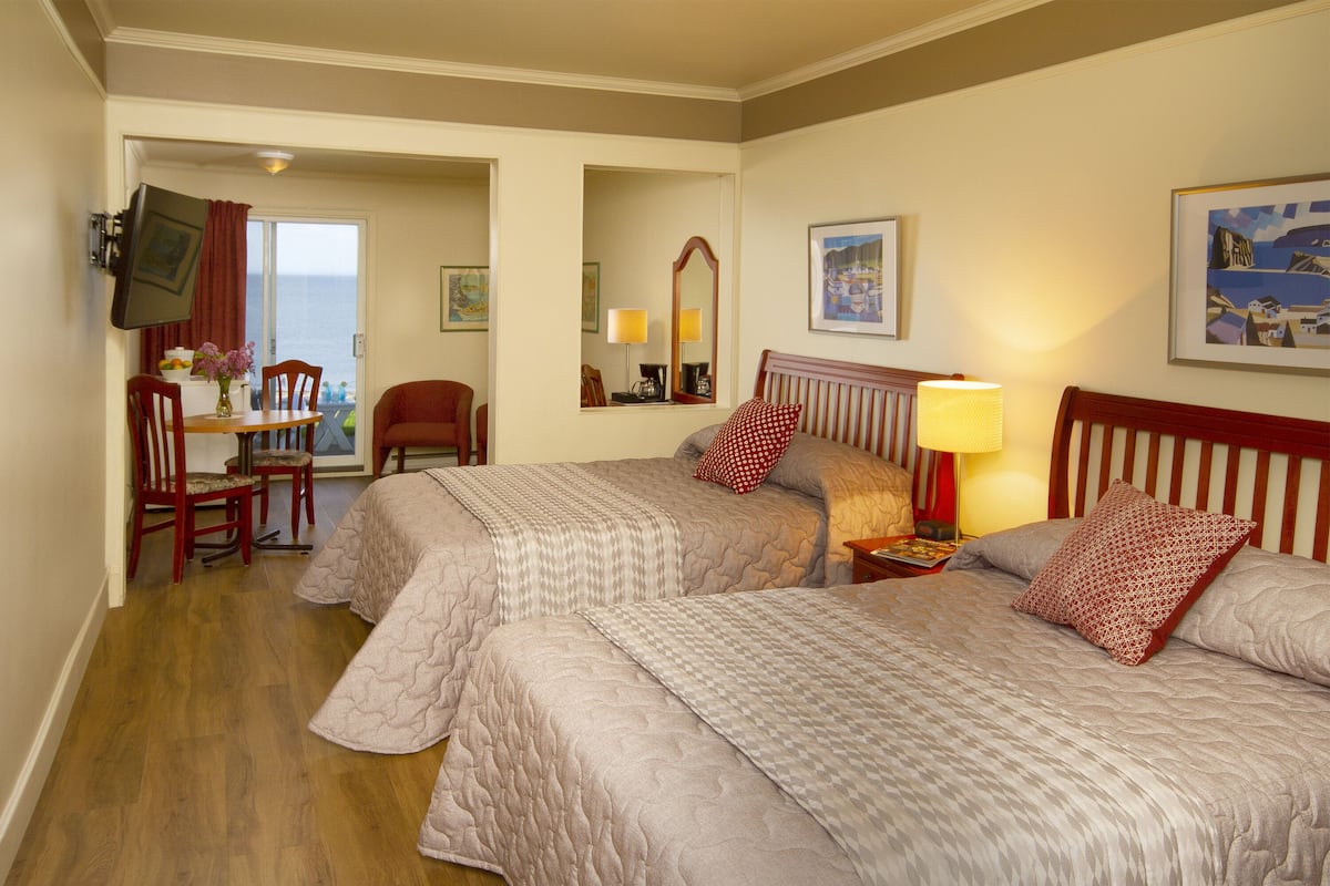 Ein Hotelzimmer mit zwei Betten, einem Esstisch mit Stühlen, einem an der Wand befestigten Fernseher und Meerblick durch eine Schiebetür.