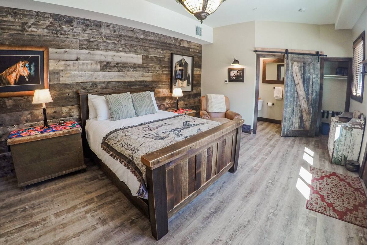 Quarto com cabeceira de madeira, uma cama grande, mesa de cabeceira com luminária, uma cadeira e banheiro visível por uma porta aberta.