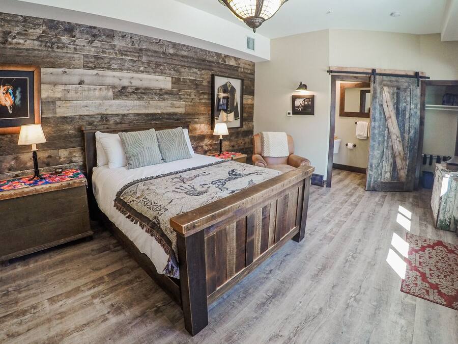 Une chambre à coucher avec une tête de lit en bois, un grand lit, une table de chevet avec une lampe, une chaise et une salle de bain visible à travers une porte ouverte.