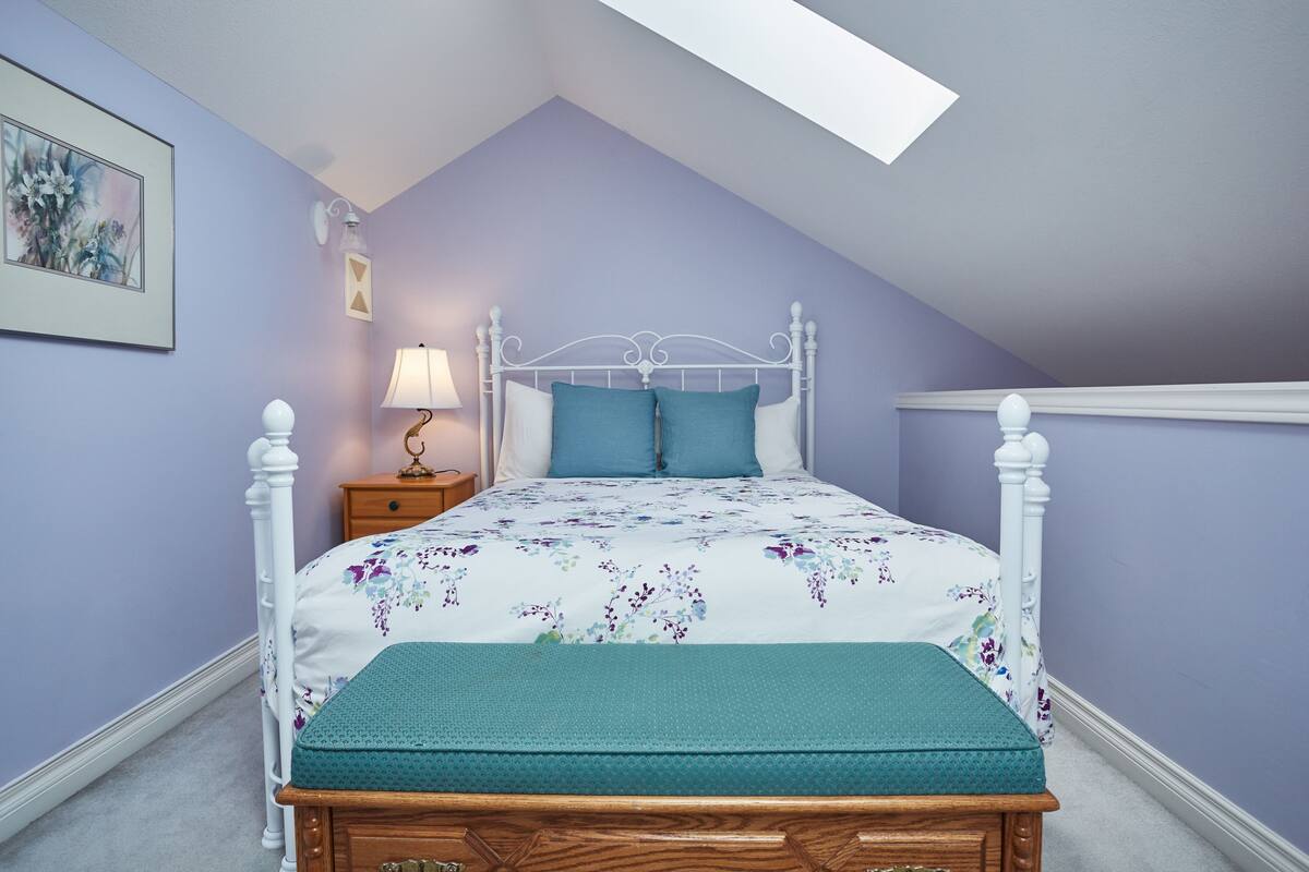 Quarto com teto inclinado, estrutura de cama de metal branco, colcha estampada com flores, um banco verde-água e mesa de cabeceira com abajur.