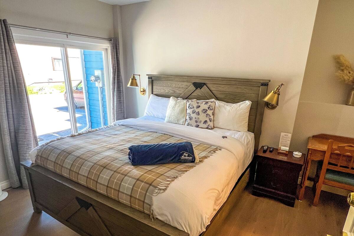 Quarto com cama de madeira, manta azul e mesa de cabeceira com abajur.