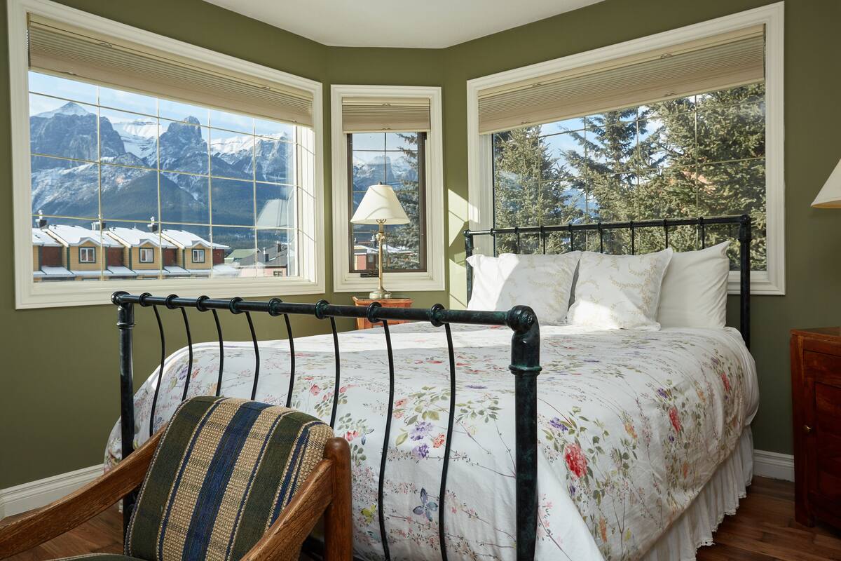 Quarto com cama, poltrona e grandes janelas com vista para as montanhas.