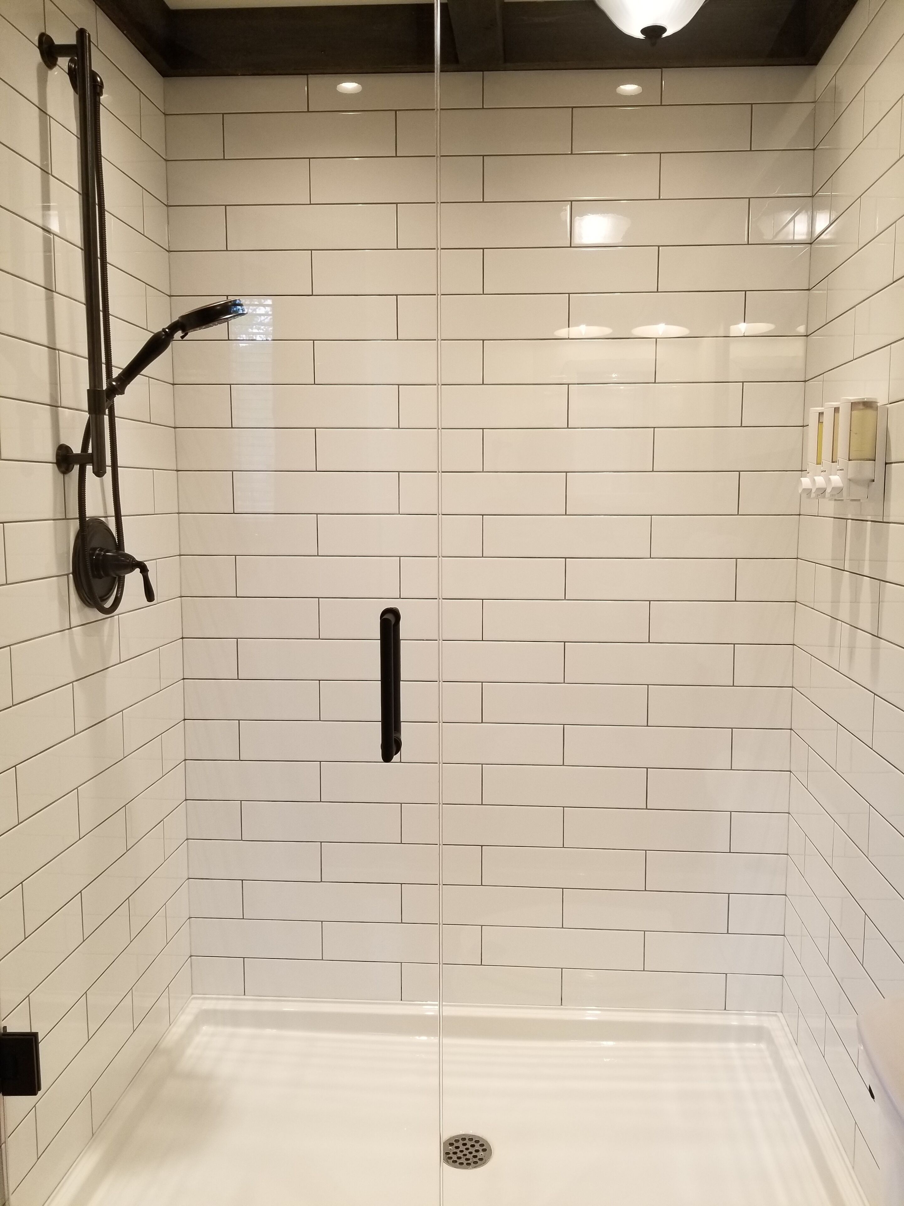 Boxe sanitário com azulejos brancos tipo metrô, chuveiro preto e maçaneta.