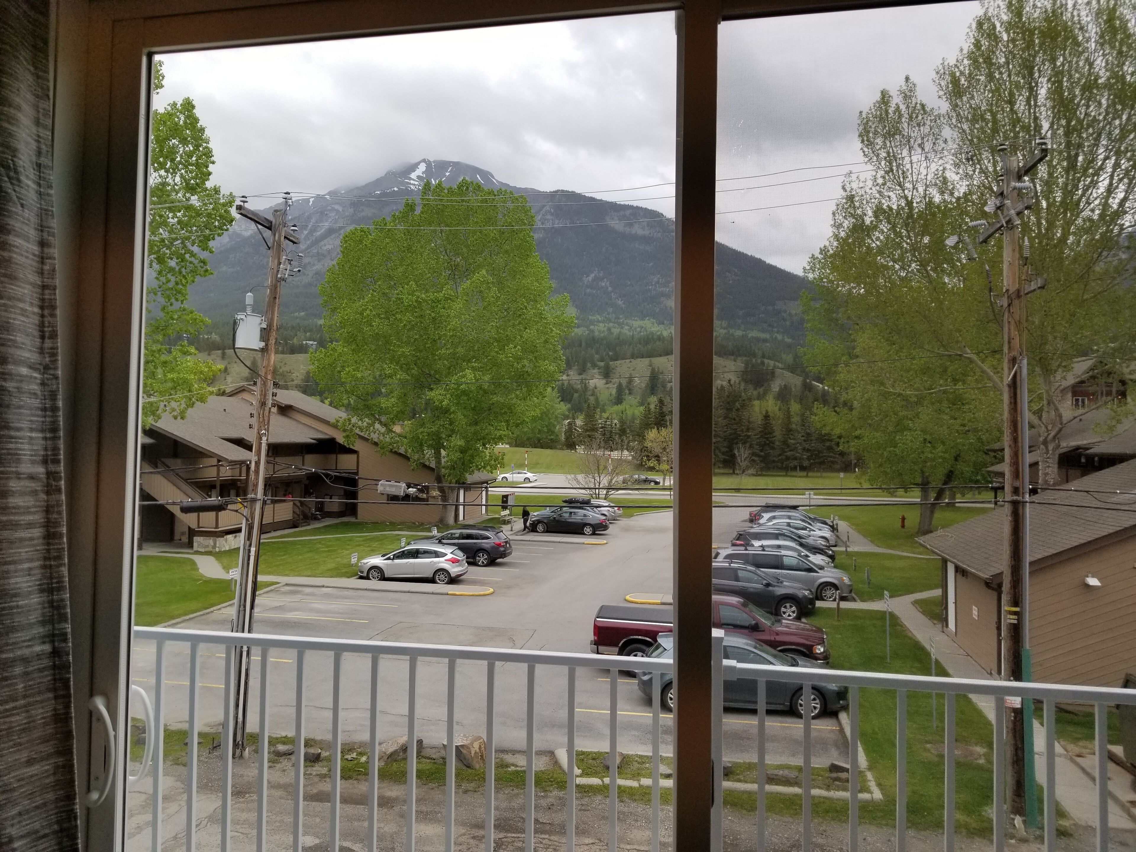 A view from a window overlooking a parking lot with several cars, a building with a sloped roof, and a mountain in the background.