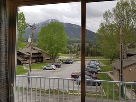 A view from a window overlooking a parking lot with several cars, a building with a sloped roof, and a mountain in the background.