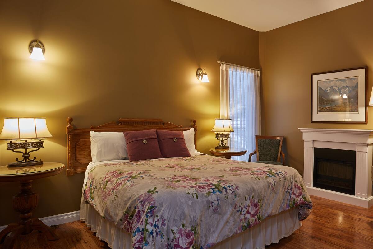Quarto com cama de madeira, colcha florida, duas luminárias de cabeceira, uma cadeira, lareira e um quadro na parede.