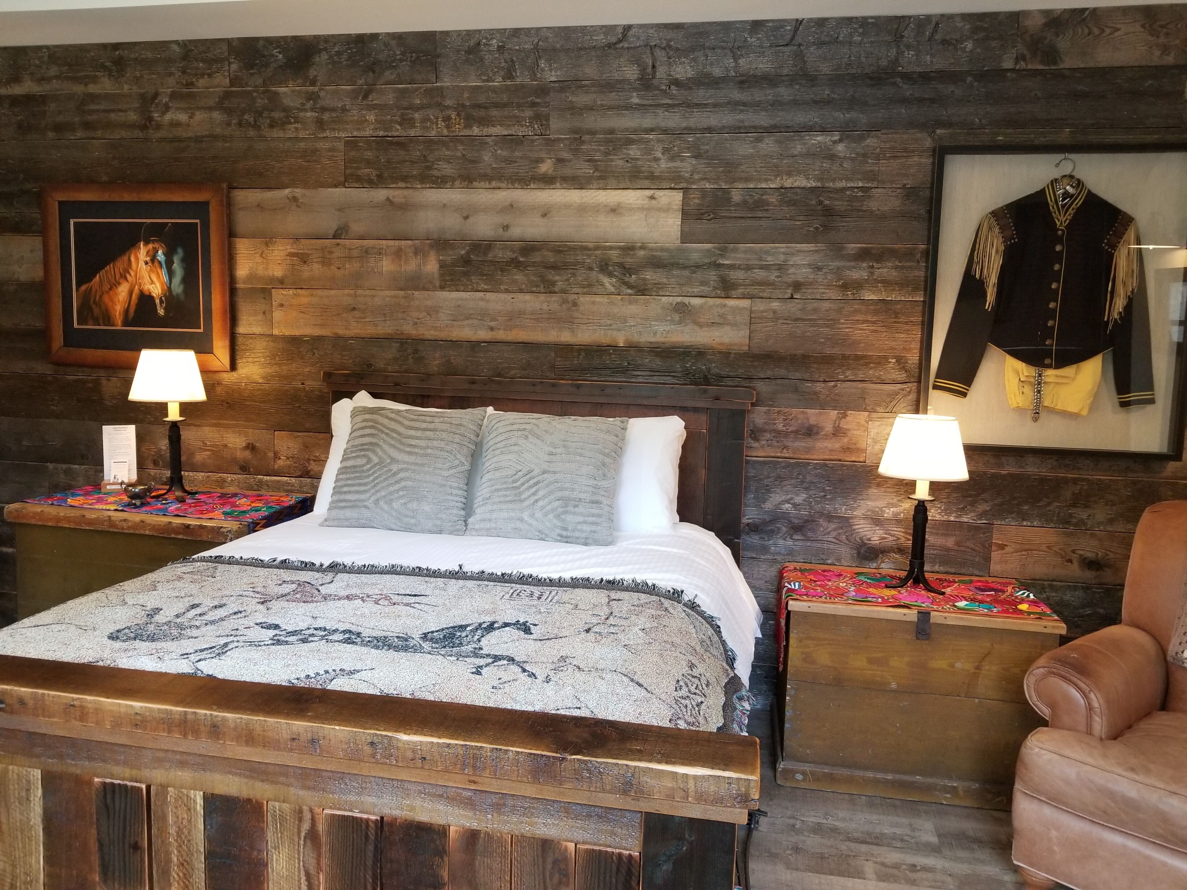 Una cama con cabecera de madera, dos mesitas de noche con lámparas, un cuadro con un caballo y uno con un uniforme militar, además de un sillón de cuero marrón.