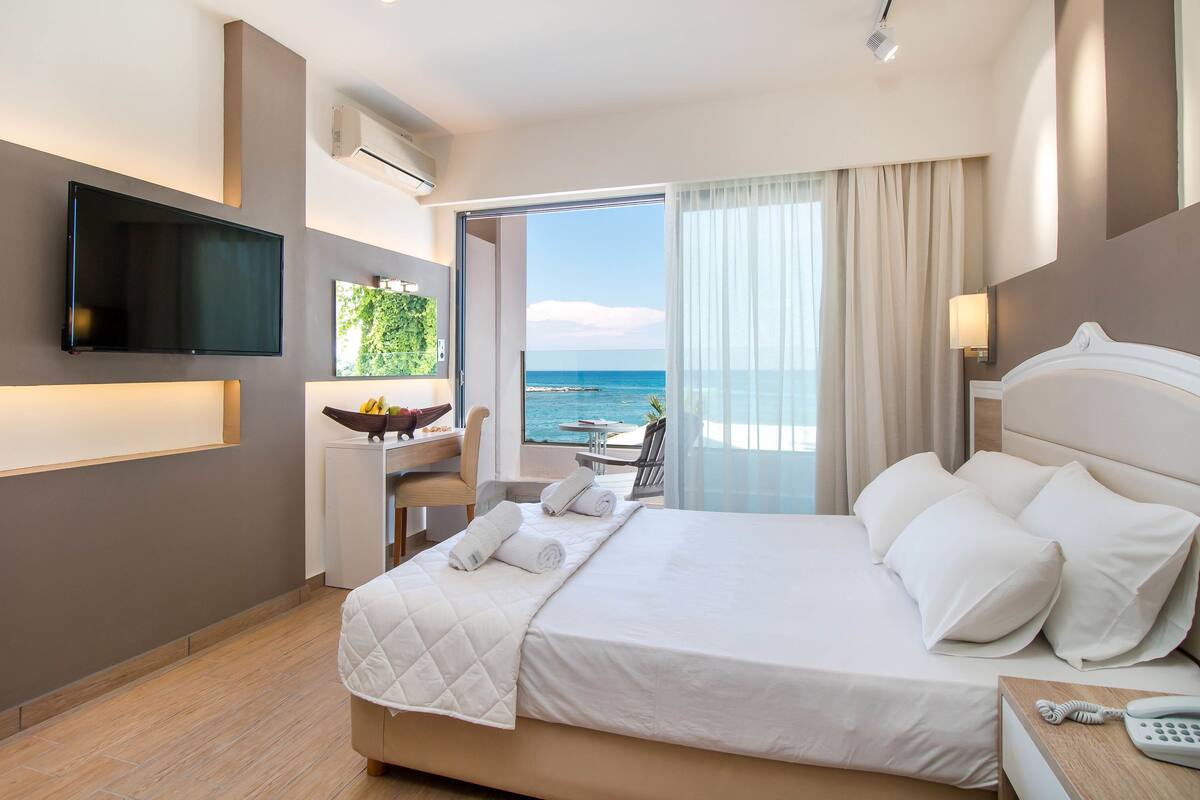 Habitación de hotel moderna con una cama grande, un televisor de pantalla plana, un escritorio pequeño con un cuenco de fruta y vista al océano.