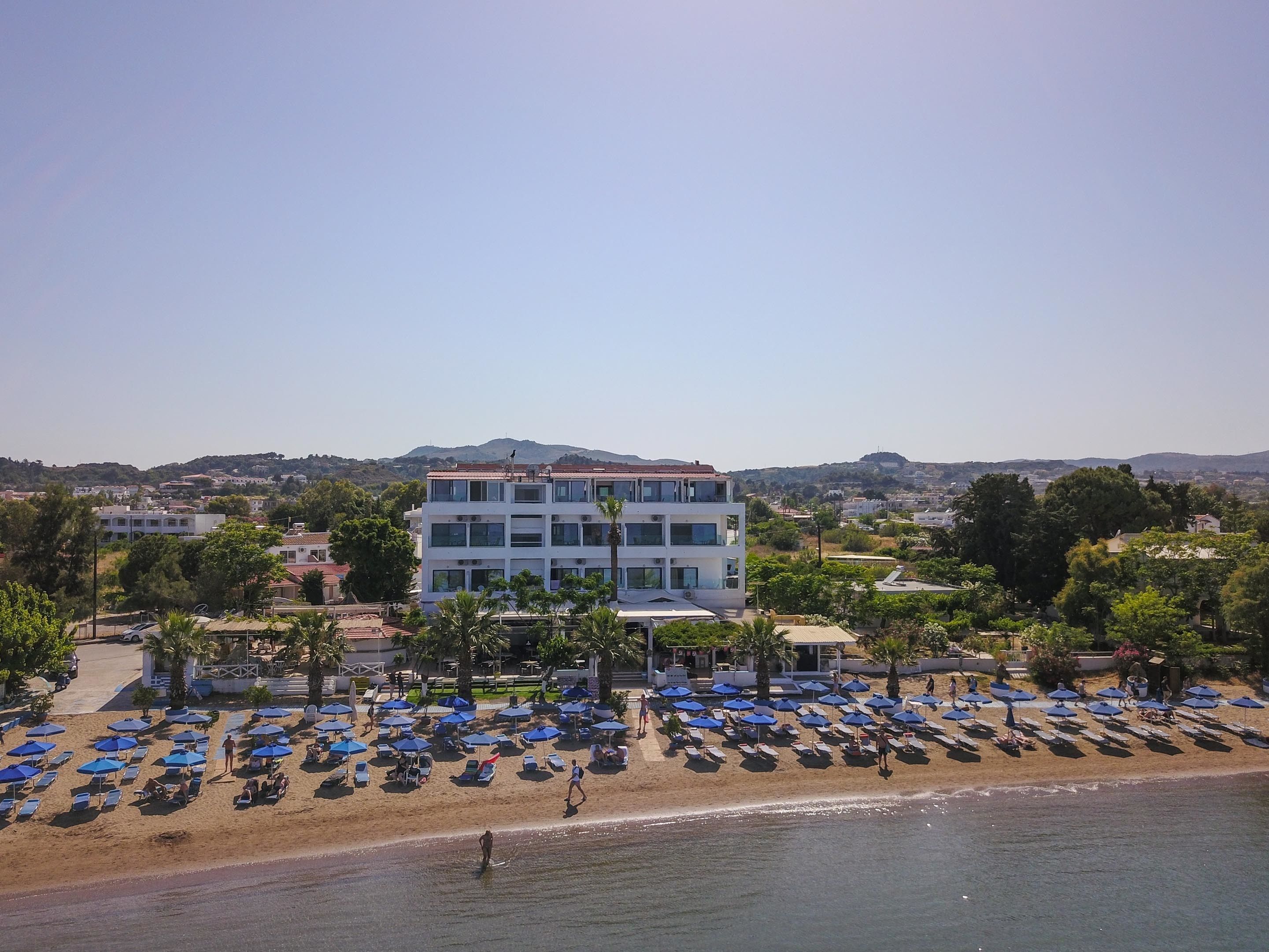 Un hotel frente a la playa con varios pisos, una playa arenosa con numerosas sombrillas y vistas del paisaje circundante, que incluye colinas y árboles.