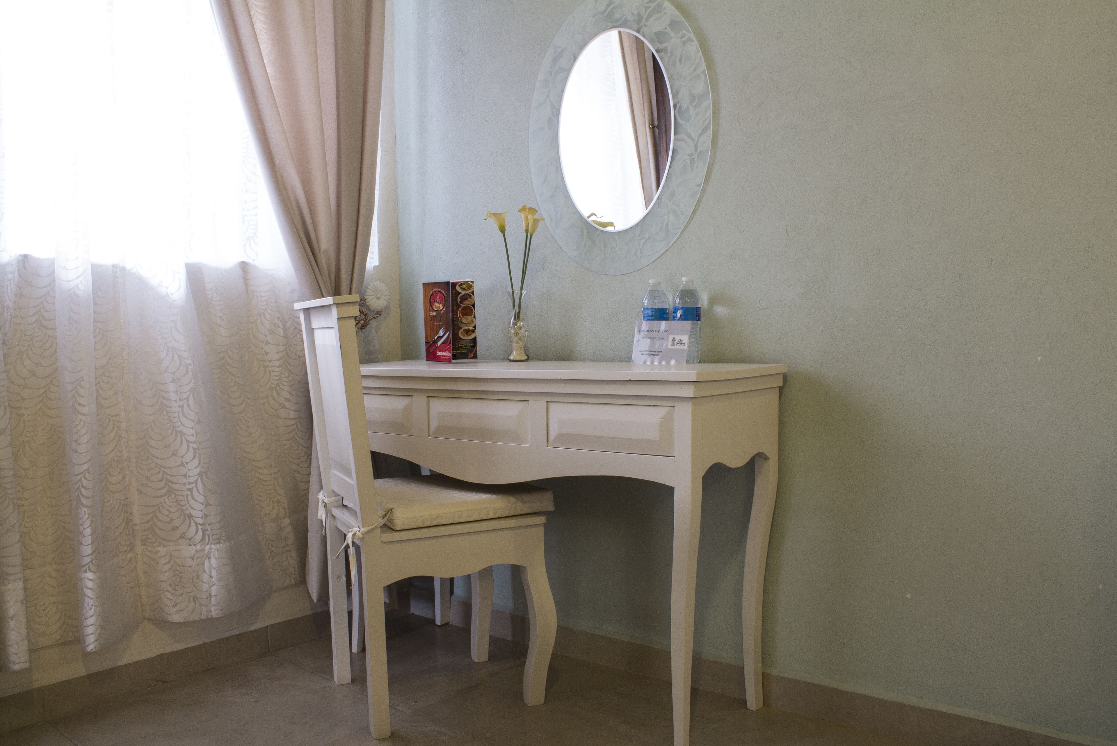 A white vanity with a mirror, a vase with flowers, and a water bottle on a dresser.