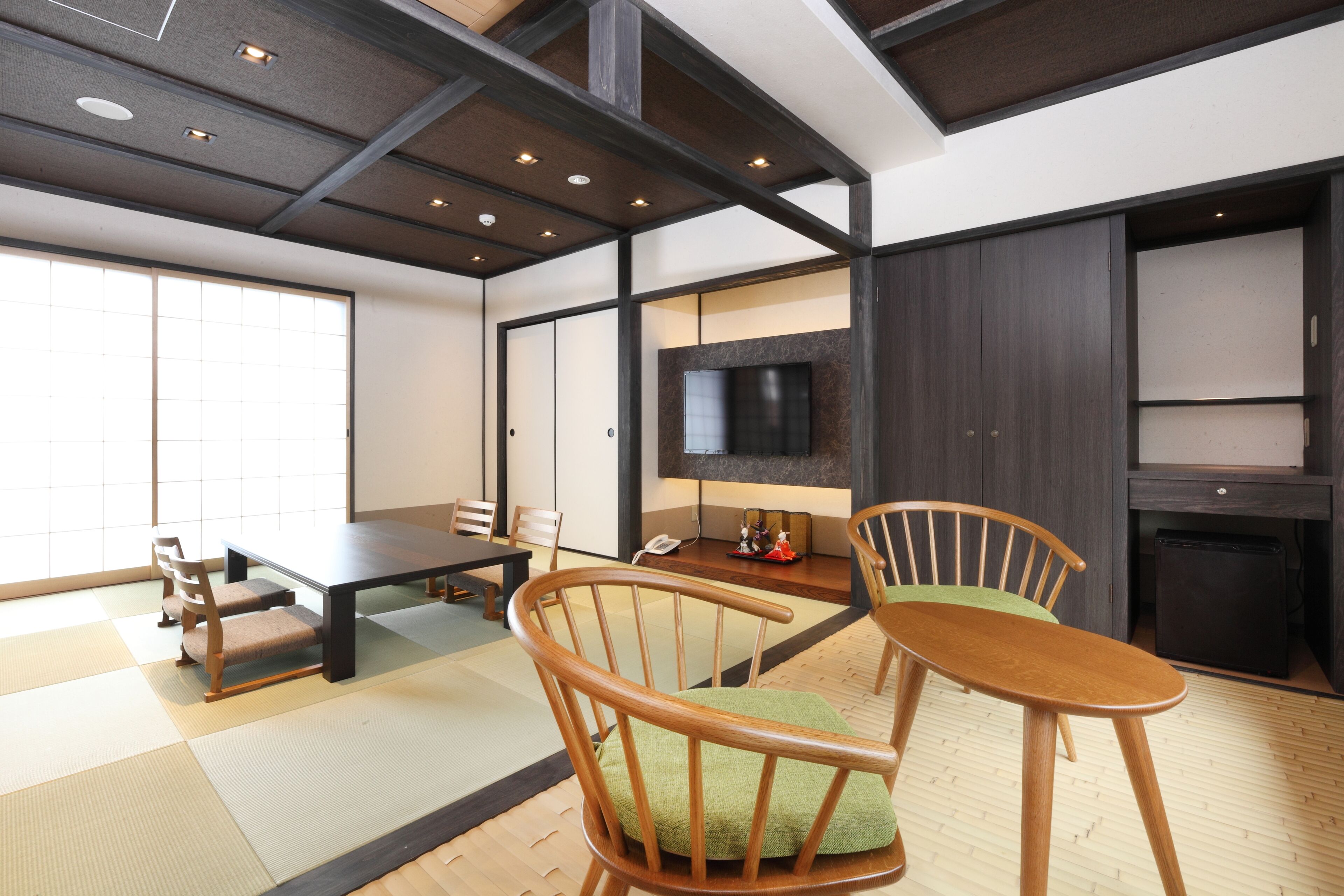Una habitación japonesa tradicional con suelo de tatami, una mesa baja, sillas de madera, un televisor y un refrigerador pequeño.