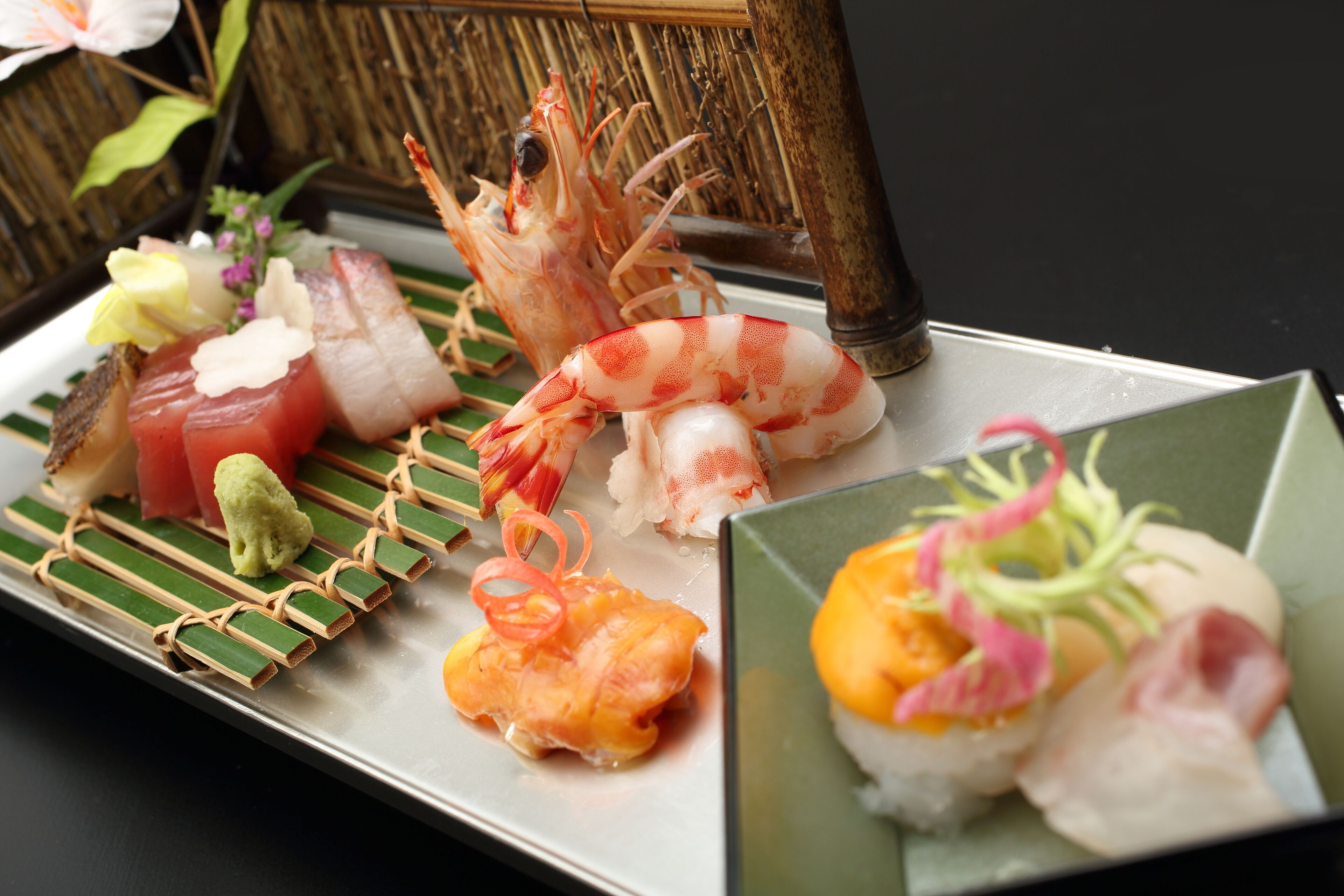 A variety of sushi dishes on a rectangular plate with a bamboo backdrop.