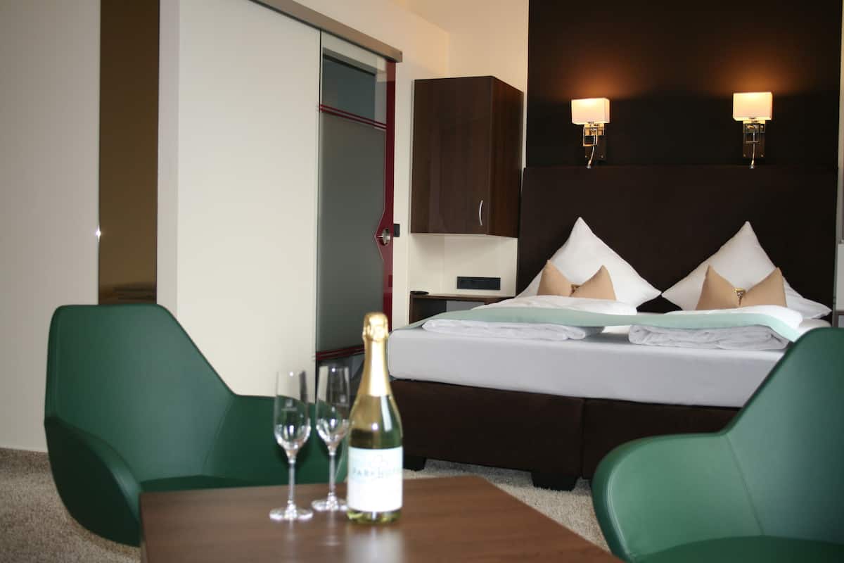 A hotel room with a bed, two green chairs, a table with a bottle of champagne and glasses, and a minibar.