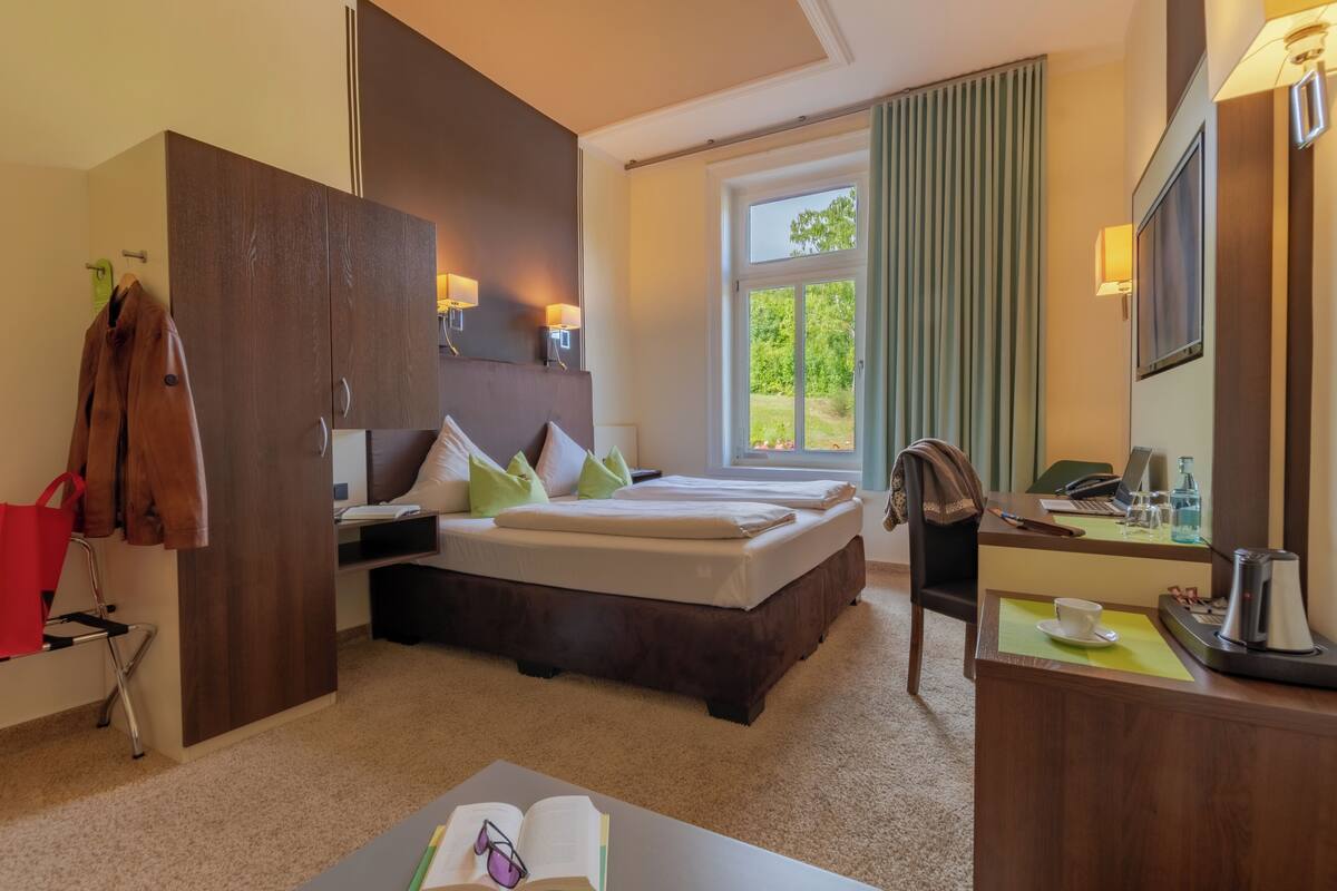 A hotel room with a bed, desk, and a window with a view of greenery.