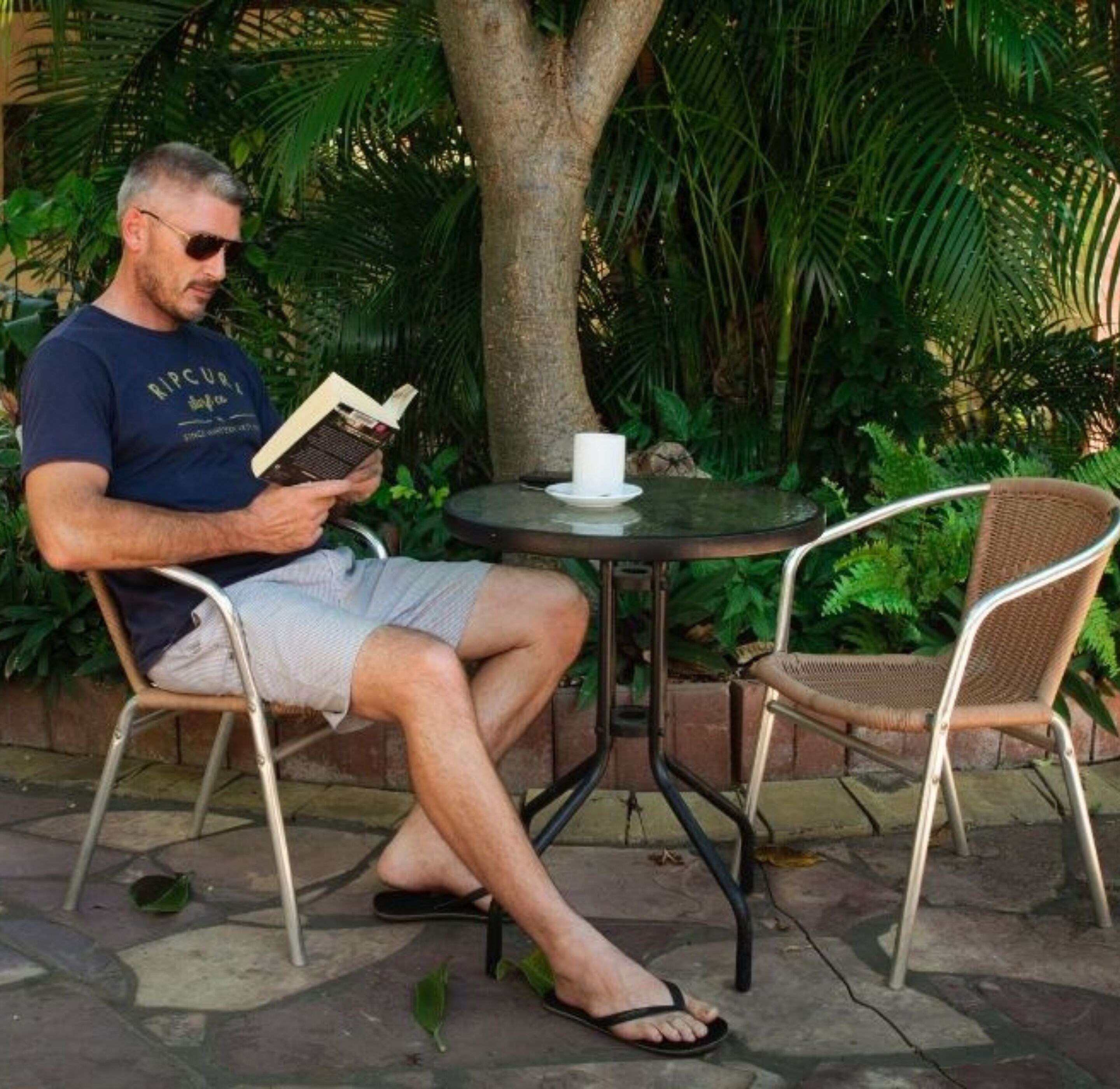 Un hombre leyendo un libro a una pequeña mesa redonda, con una taza blanca a su lado, sentado en una silla de mimbre en un jardín.