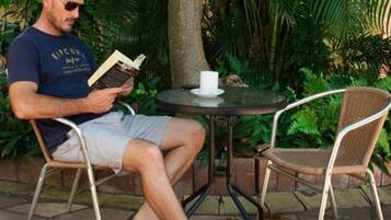 Un hombre leyendo un libro a una pequeña mesa redonda, con una taza blanca a su lado, sentado en una silla de mimbre en un jardín.