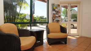 Una habitación con sillas de mimbre y una mesa, una ventana con vista a palmeras, y una puerta con varios letreros.