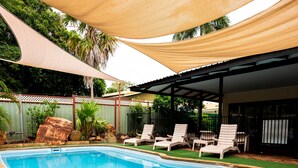 Una piscina rectangular con borde de azulejos, rodeada de césped y un patio sombreado con sillas de descanso blancas.