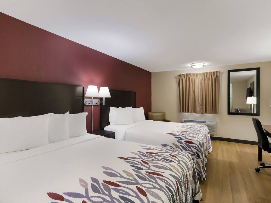 A hotel room with a large bed, a desk, a chair, a nightstand, a lamp, a mirror, and a window with curtains.