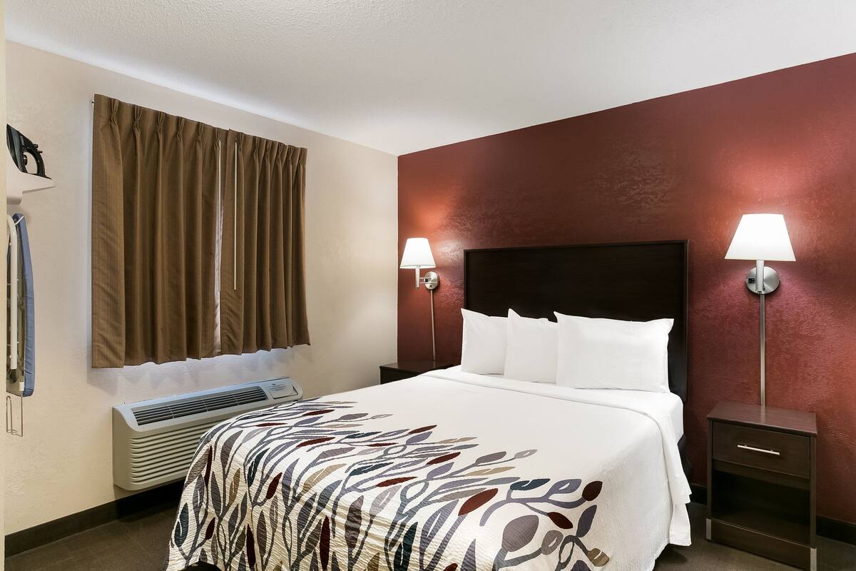 Quarto de hotel com cama, duas luminárias de cabeceira, janela com cortinas, ar condicionado e mesa de cabeceira.