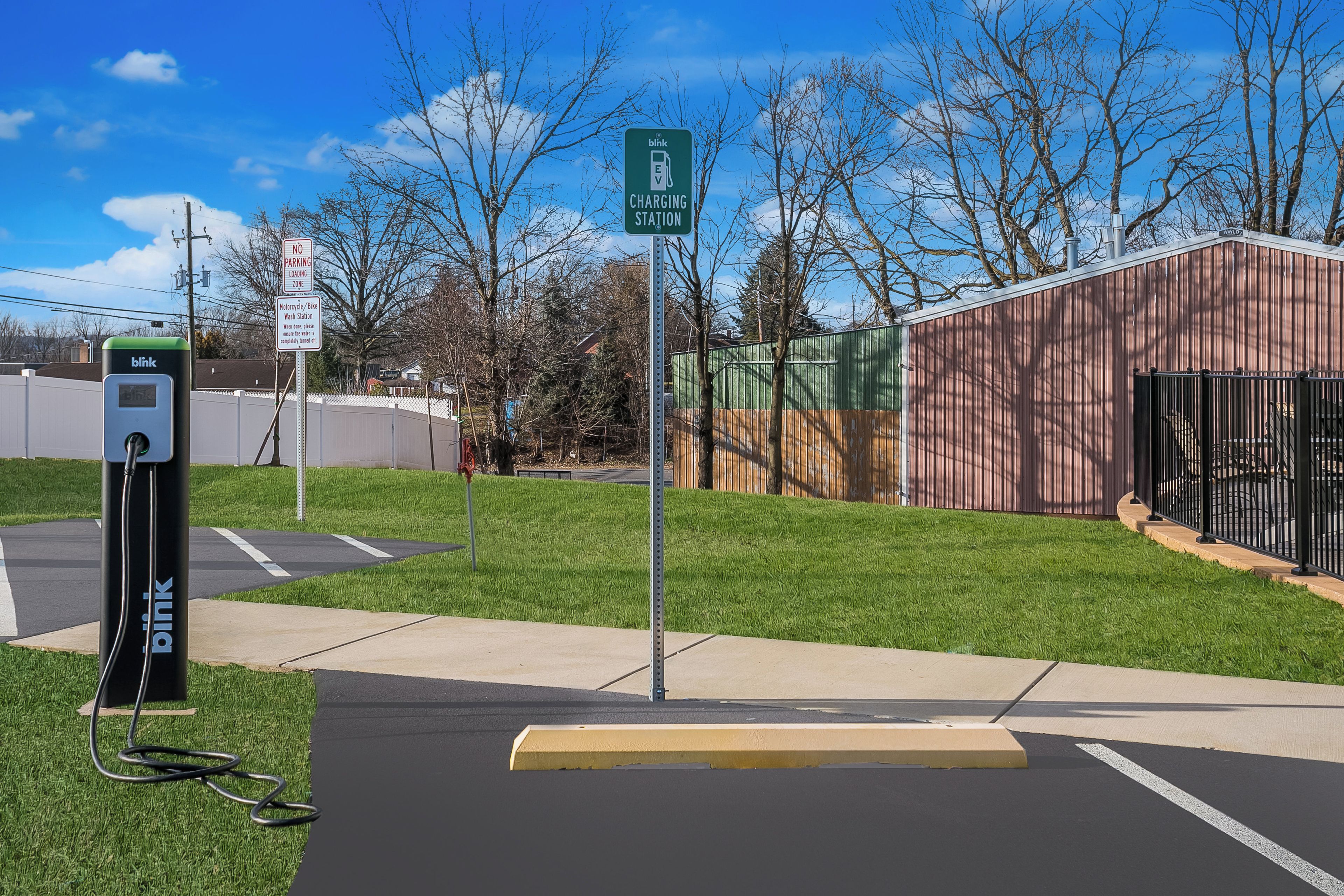 A Blink EV charging station with a no parking zone sign, a motorcycle wash station, and a fenced area with a building in the background.