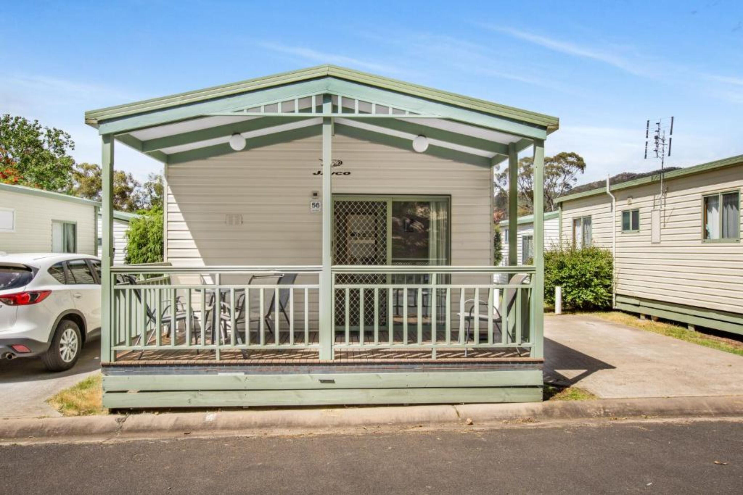 Une petite maison mobile d’un seul étage, dotée d’un toit végétalisé et d’un revêtement extérieur blanc. Elle comprend une véranda couverte avec une rambarde en métal et une petite fenêtre.