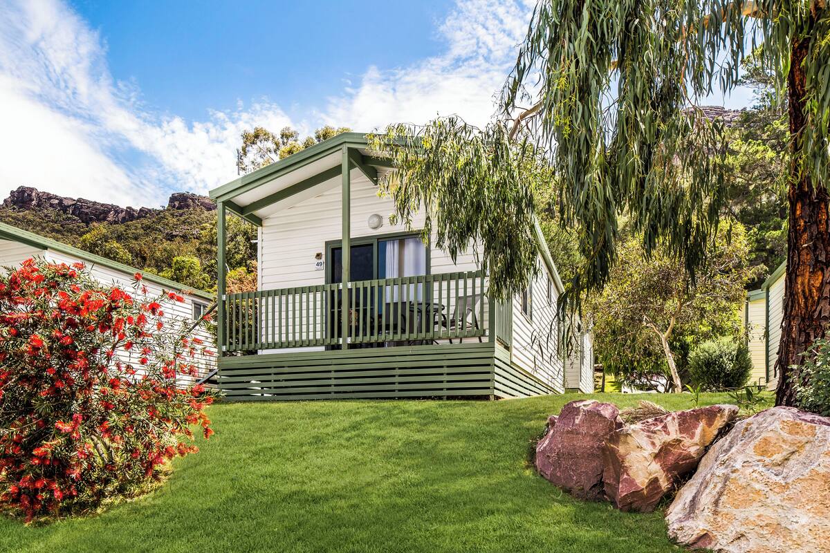 Een gelijkvloers huis met een veranda, omgeven door een verzorgd gazon en grote rotsen.