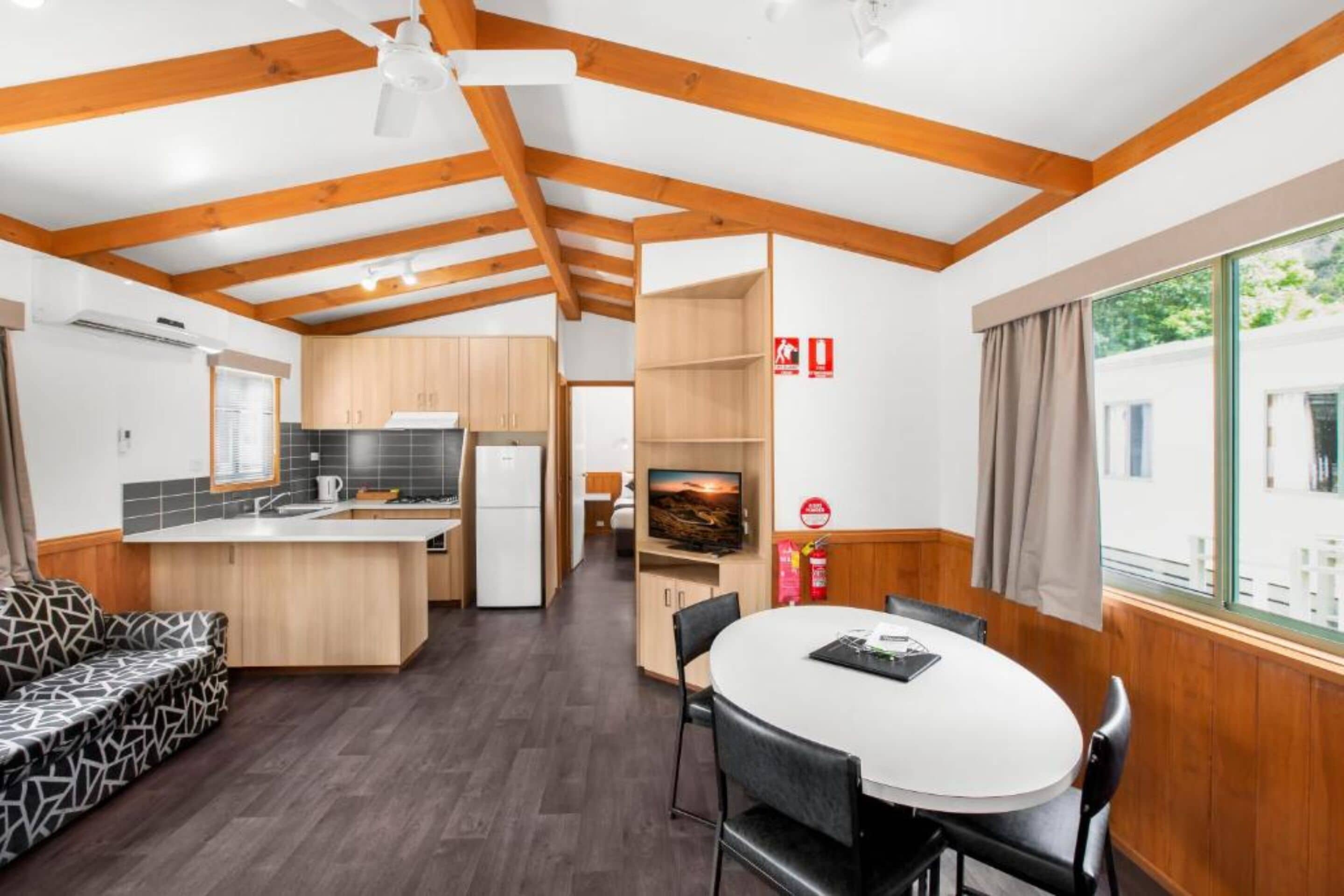 A modern kitchen with wooden beams, a dining area with a round table, and a TV in the background.
