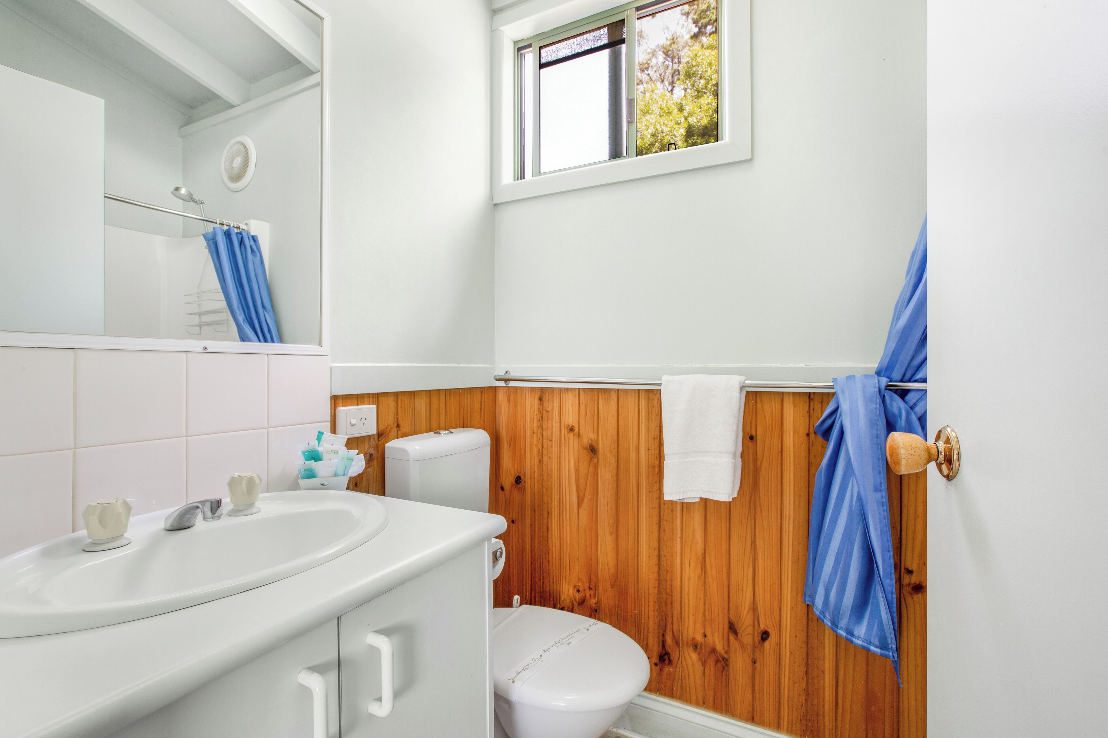 Une salle de bain avec un mur lambrissé en bois, un lavabo blanc, des toilettes et une fenêtre donnant sur des arbres.