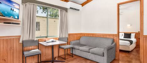 Una habitación acogedora con techo de madera, un sofá gris, una mesita con sillas, un televisor en la pared y una cama en un rincón.