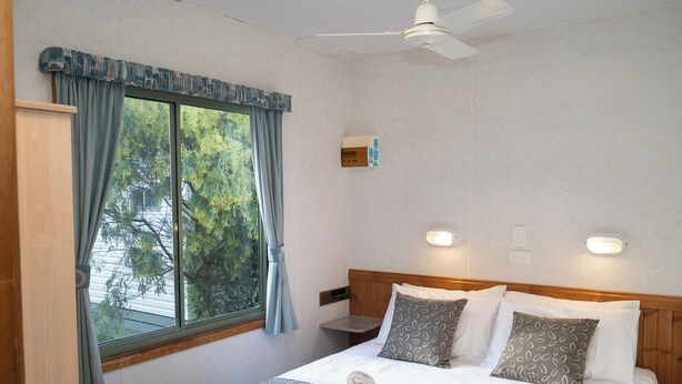 A hotel room with a bed, a nightstand, a ceiling fan, and a window with curtains. - Discovery Parks - Halls Gap (Halls Gap)