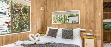 A bedroom with a wooden paneled wall, a bed with a patterned bedspread, two pillows, a bedside table, a nightstand, and a window with a view of greenery.